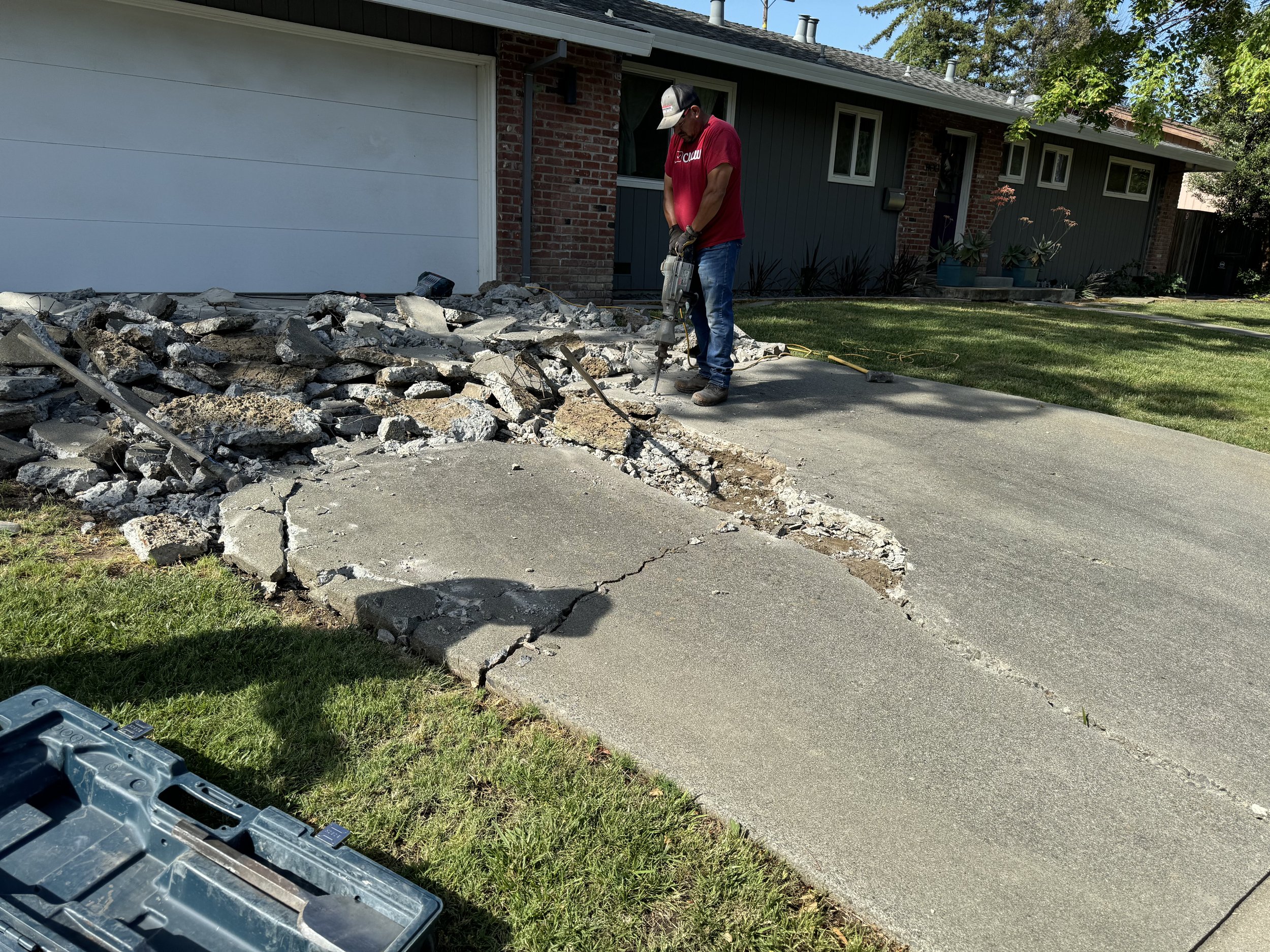 A worker in a red shirt and cap breaking a concrete driveway with a jackhammer during home driveway repair.