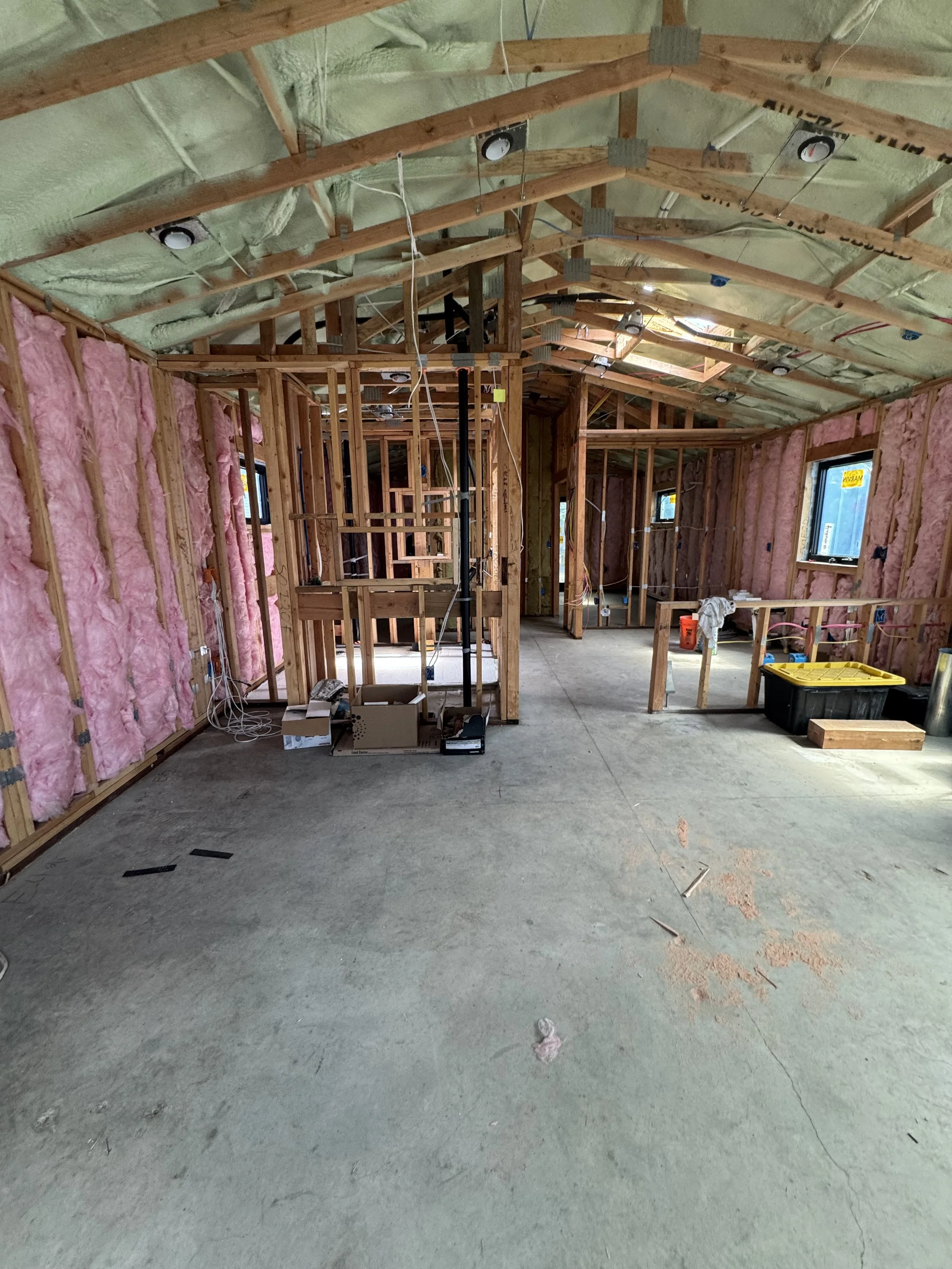 Interior of a house under construction with exposed wooden framing, pink insulation on walls, and hanging electrical wires. The floor is concrete, and there are several windows with blue sky visible outside.