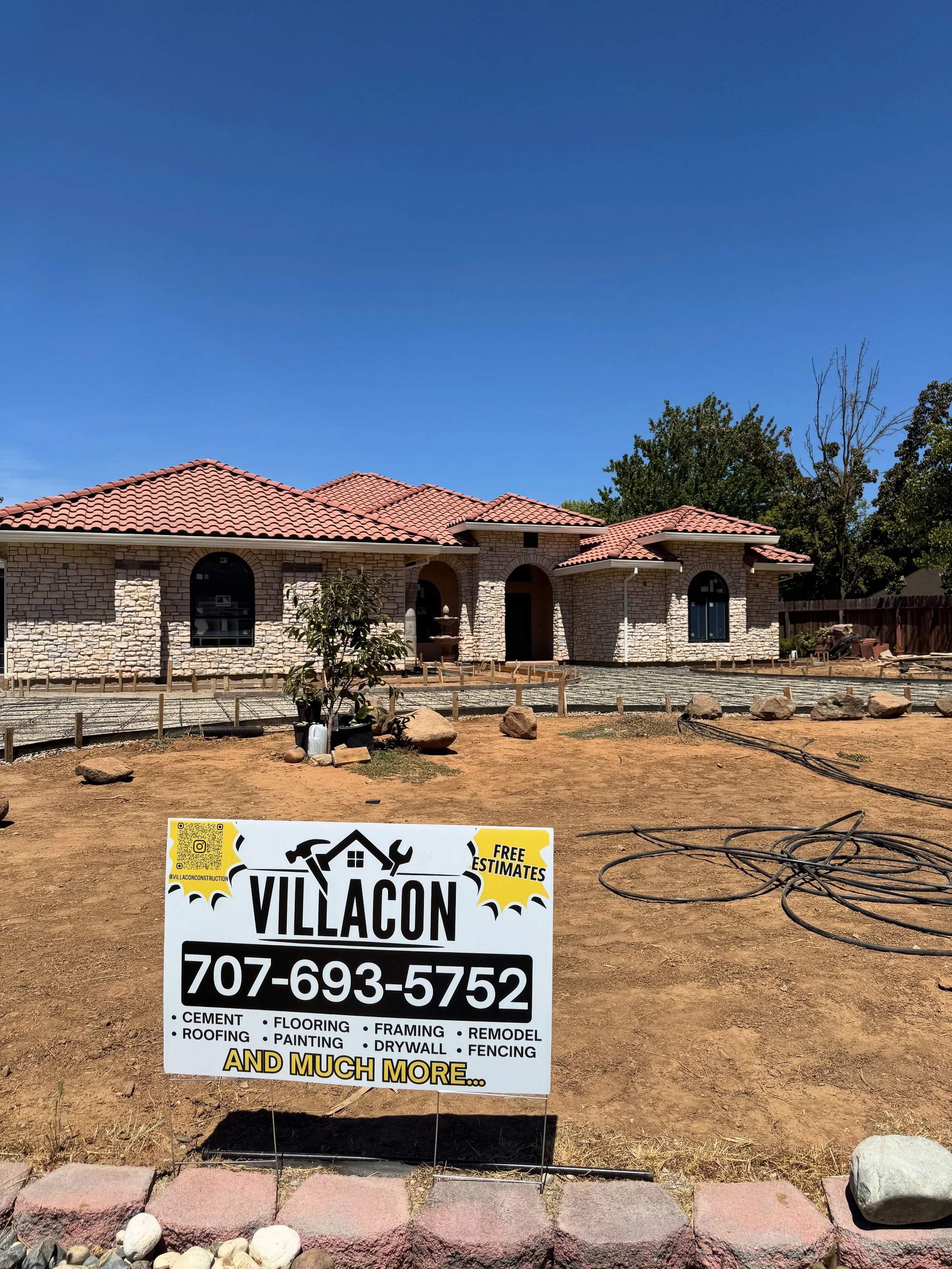 A house under construction with a red tile roof and stone exterior walls. There is a sign in the foreground advertising Villacon construction services, including cement, roofing, flooring, framing, remodeling, painting, drywall, and fencing, with a phone number for free estimates.
