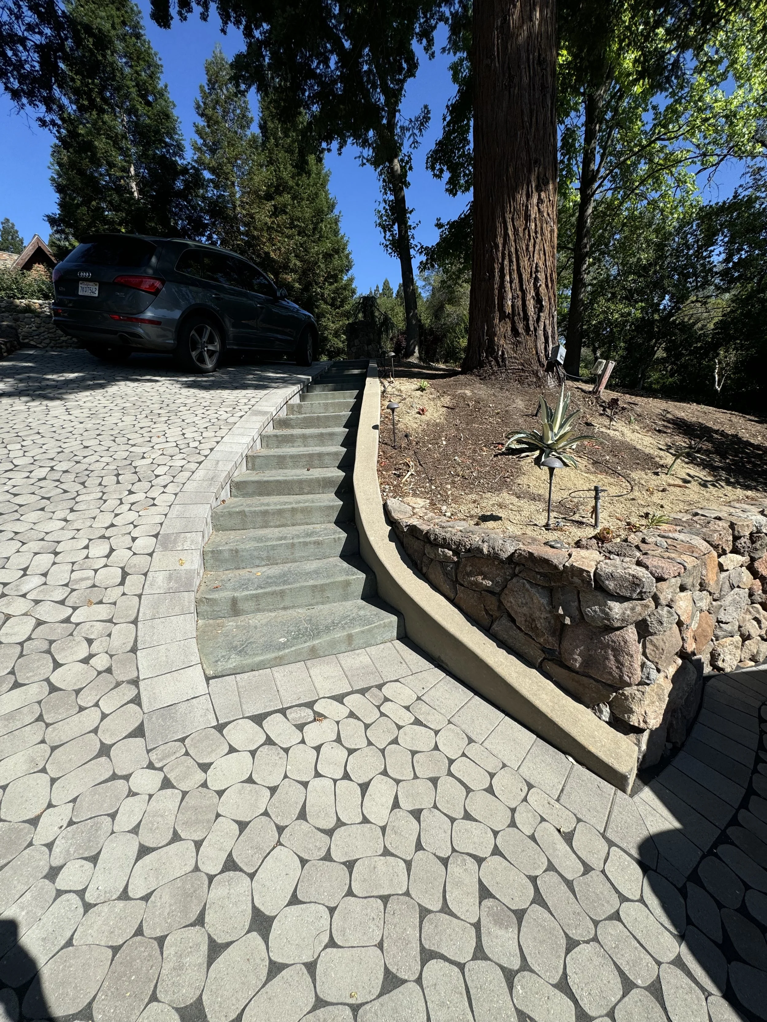 A curved outdoor staircase with concrete steps and a stone retaining wall next to a large tree, with a car parked on a patterned stone driveway in the background.