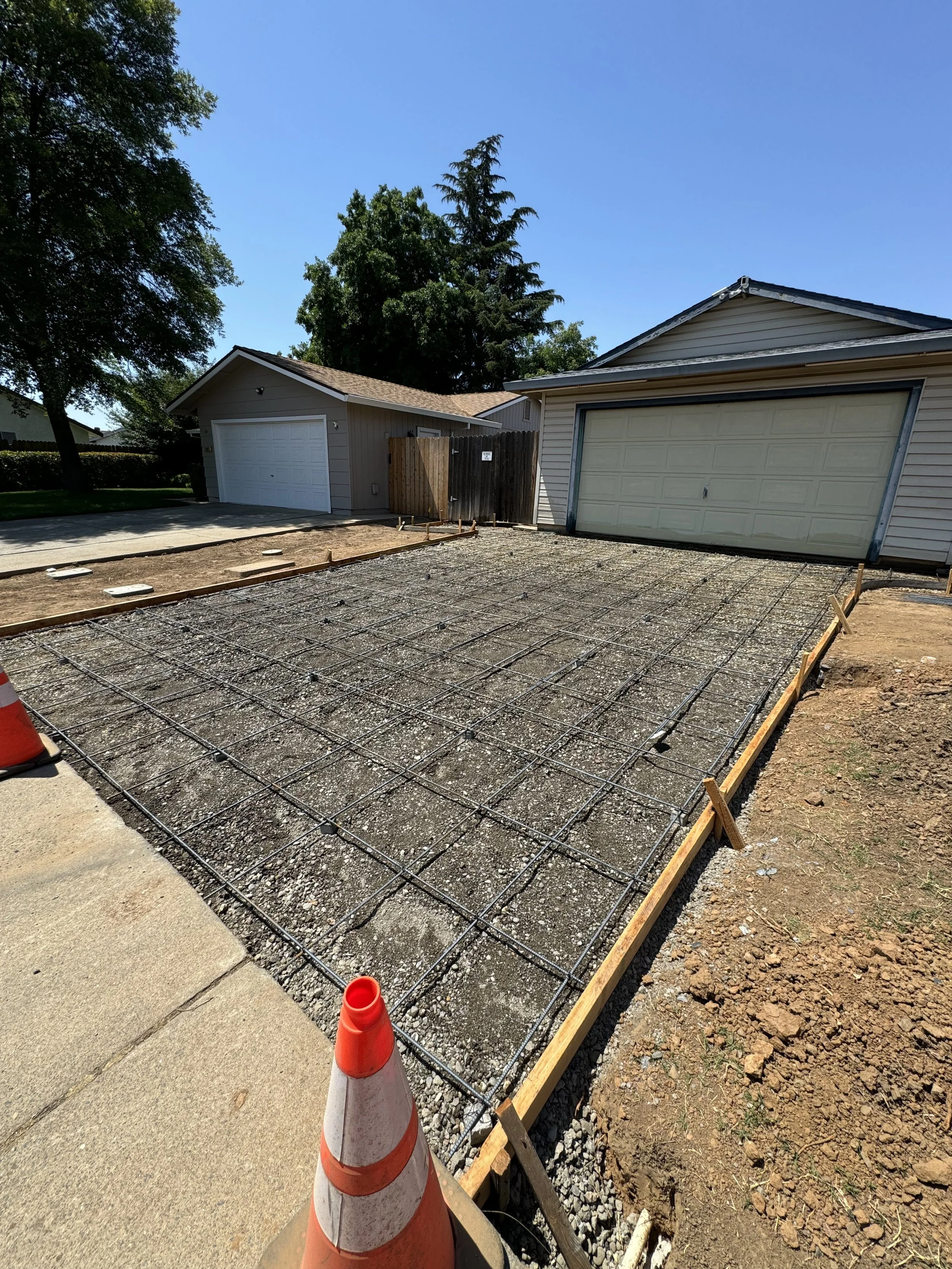 A driveway under construction with rebar grid and gravel, two garage doors, and orange construction cones.