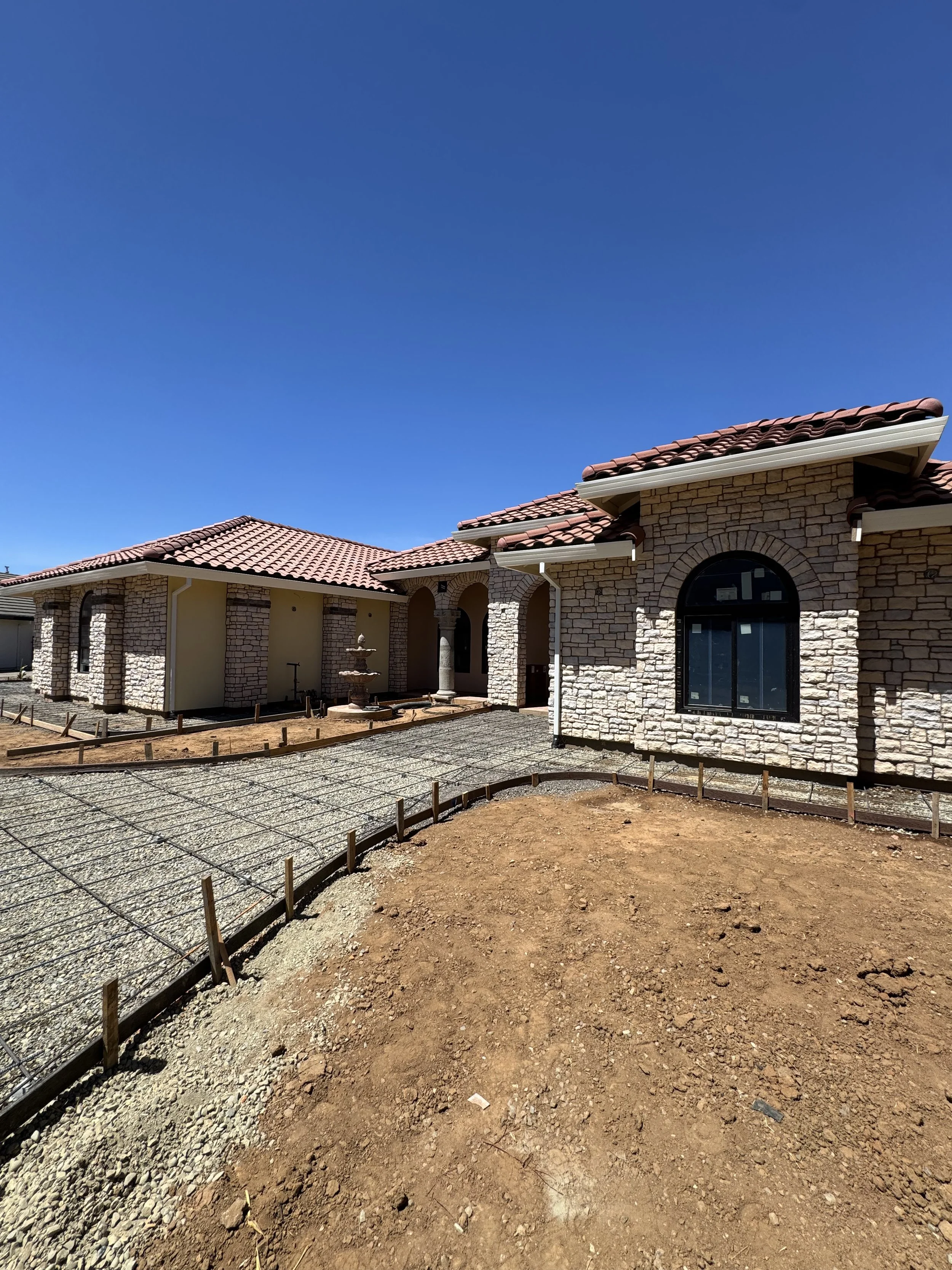 Newly constructed house with stone exterior walls, red tile roof, and fresh landscaping, including gravel walkways and bare soil yard, under clear blue sky.