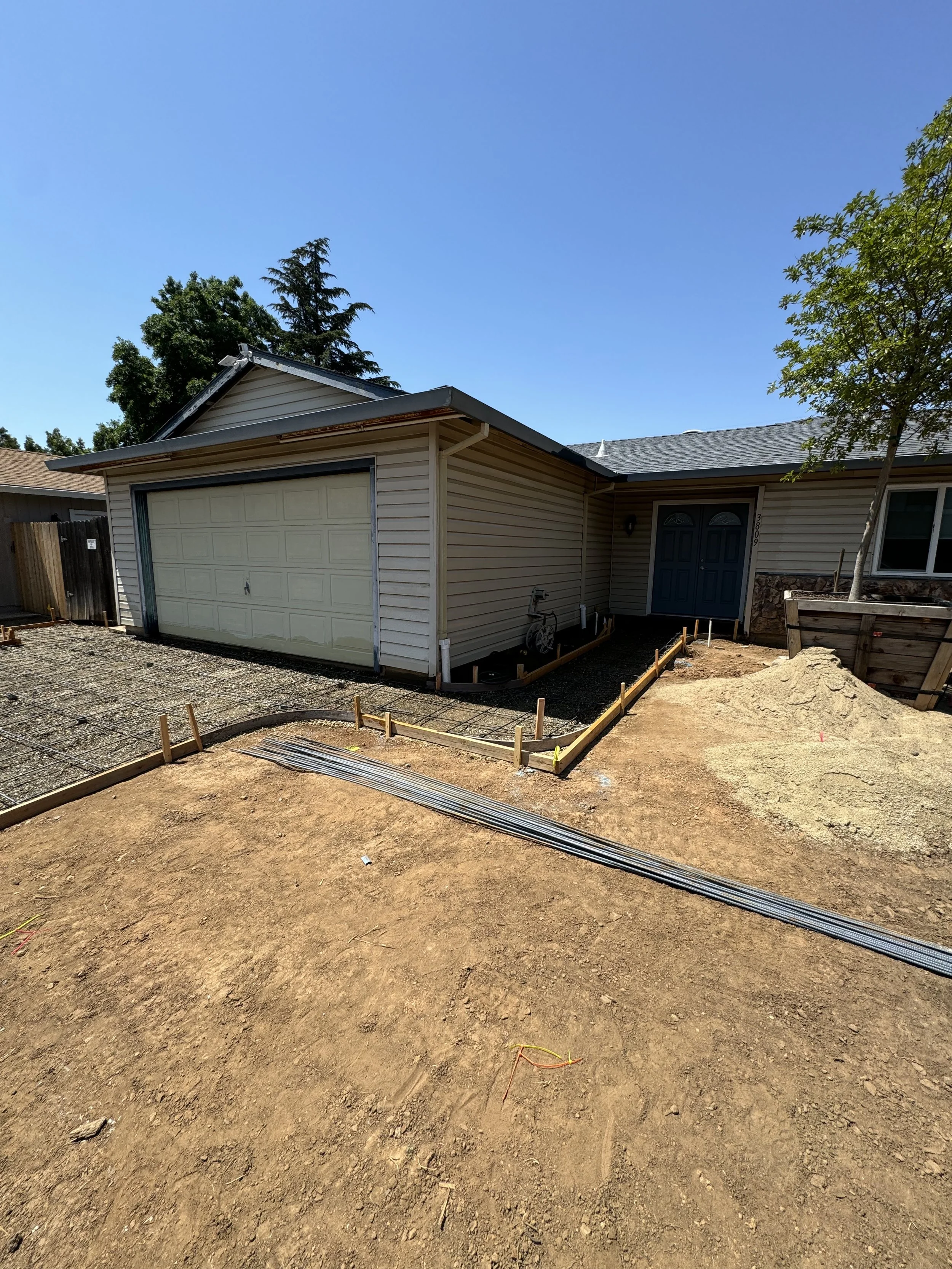 Front yard of a house under construction with exposed dirt and wooden framing, next to a house with a concrete garage door and blue front door, and a pile of dirt on the right.