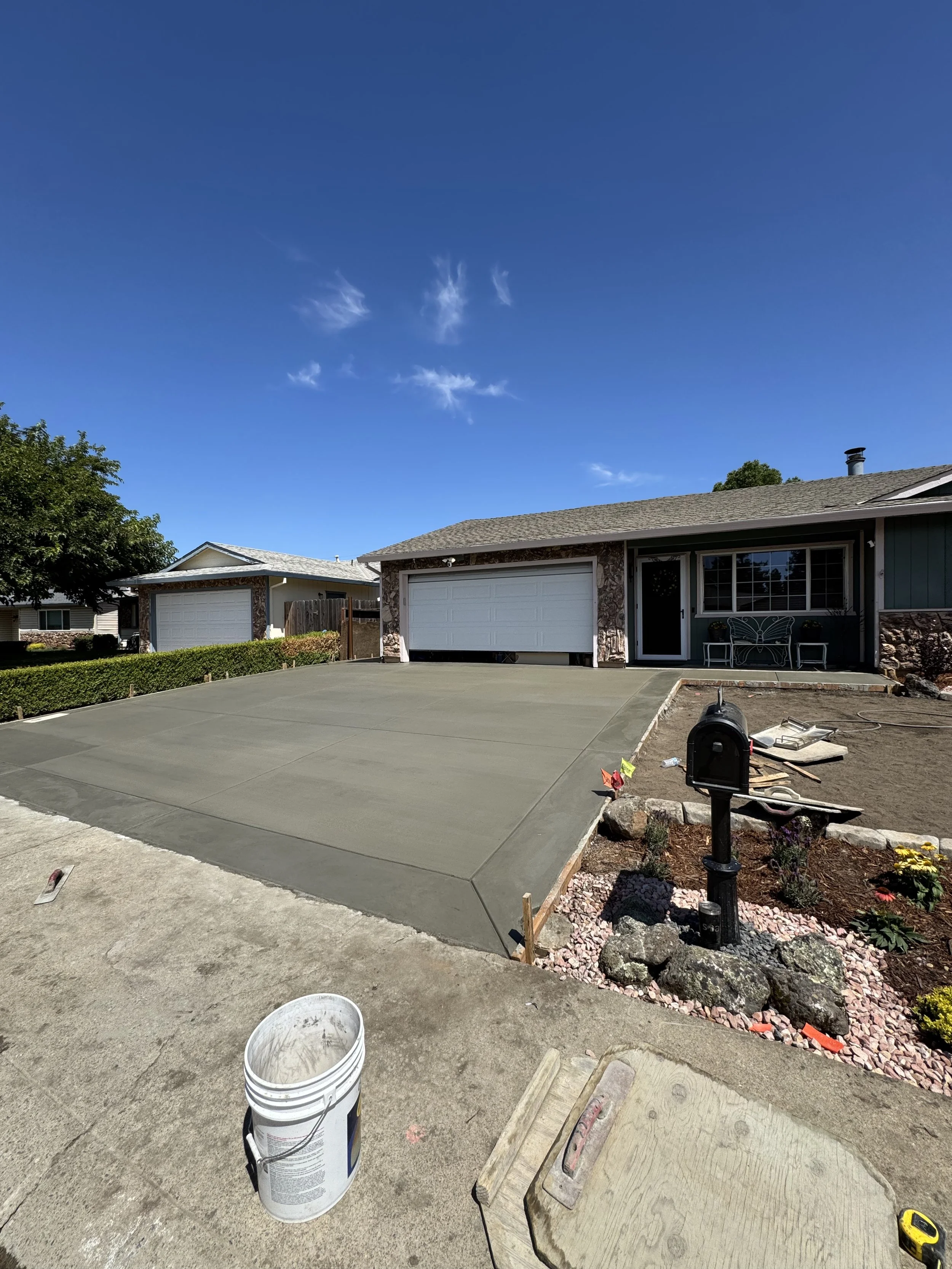 Recently paved concrete driveway in front of a house with a two-car garage, a mailbox, and a small garden with flowers; a bucket and construction materials are nearby, under a clear blue sky.
