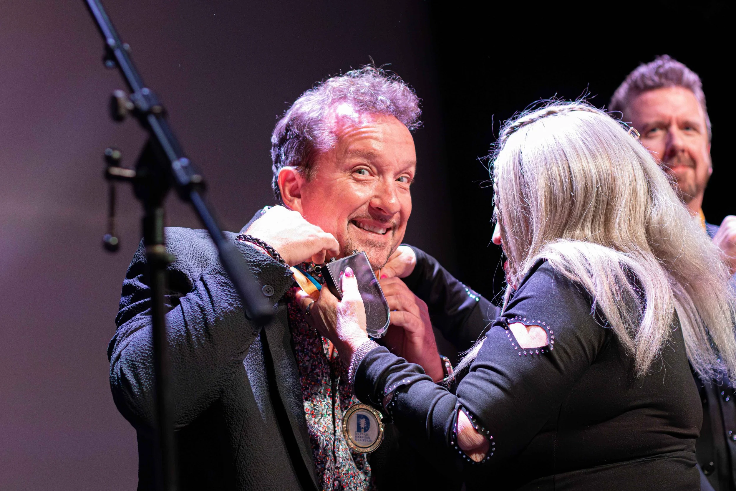 A man with curly hair smiling as a woman places a medal around his neck, with a third man smiling in the background, on a stage.