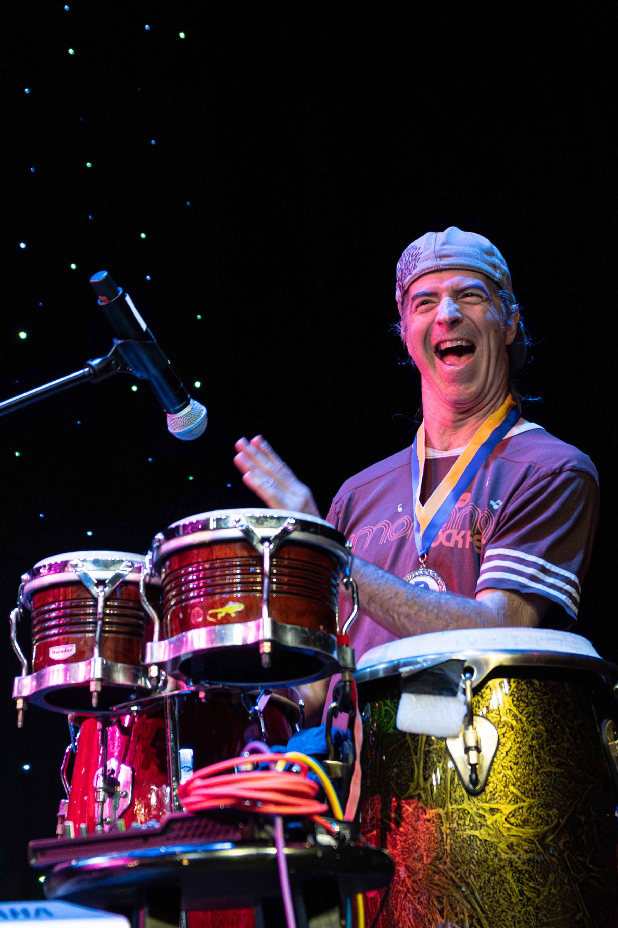 A joyful man playing percussion instruments on stage, wearing a grey cap and medal around his neck, with a microphone nearby and a dark background with small lights.