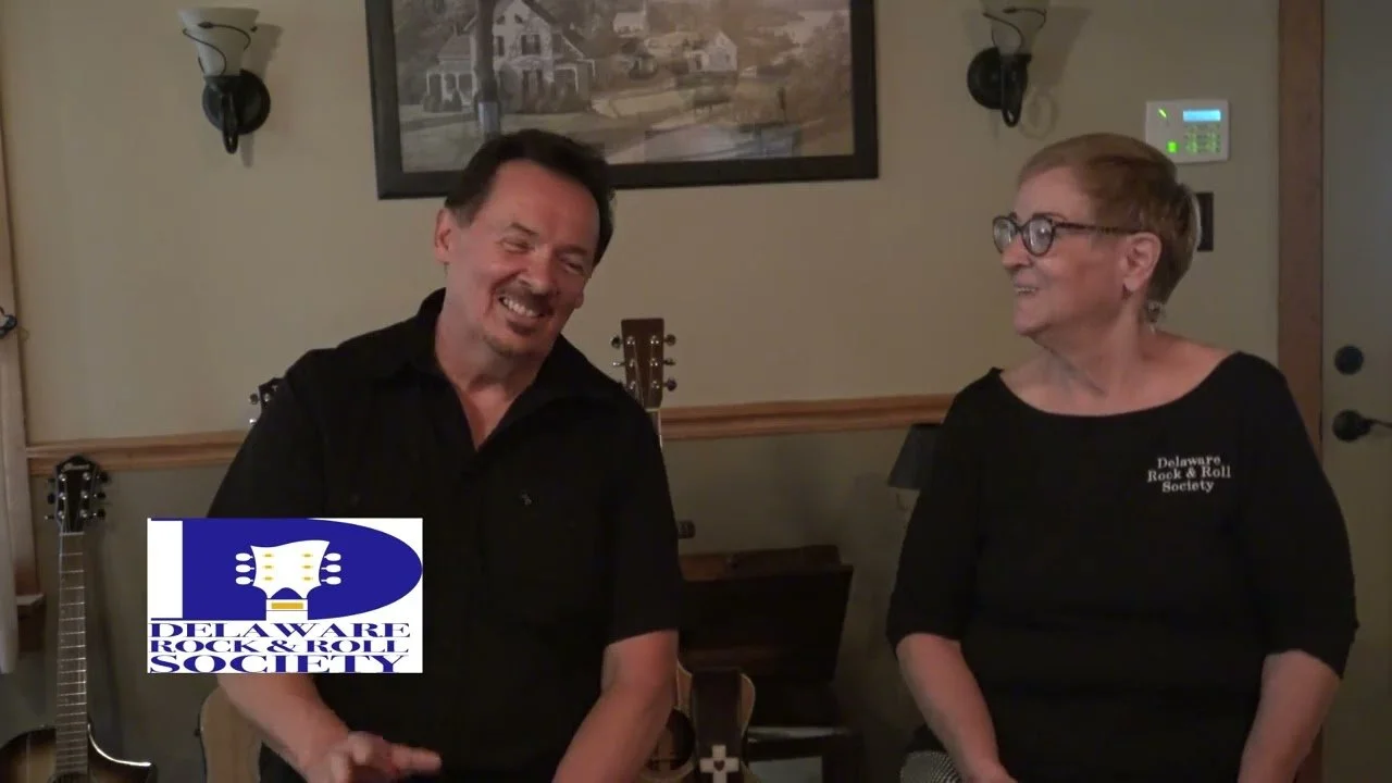 Two people, a man and a woman, are standing in a room with musical instruments, smiling and laughing. The woman is wearing glasses and a black shirt that reads 'Delaware Rock & Roll Society.' There is a large framed picture on the wall behind them and a logo for Delaware Rock & Roll Society in the bottom left corner.