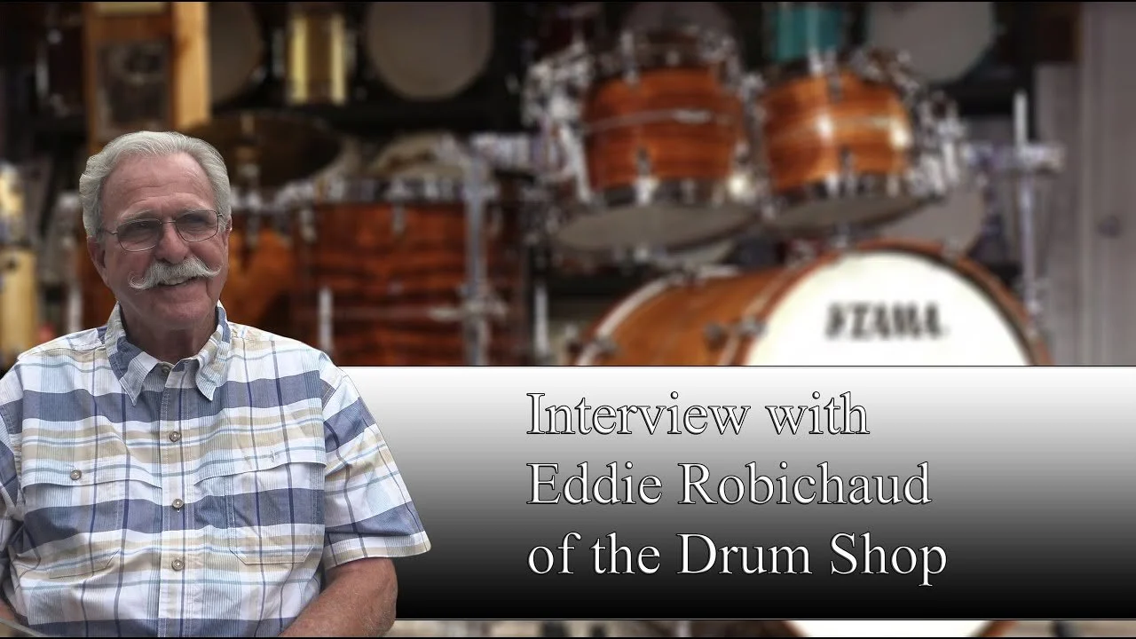 Interview with Eddie Robichaud of the Drum Shop, featuring a man with white hair and glasses, wearing a plaid button-up shirt, standing in front of a bass drum set.