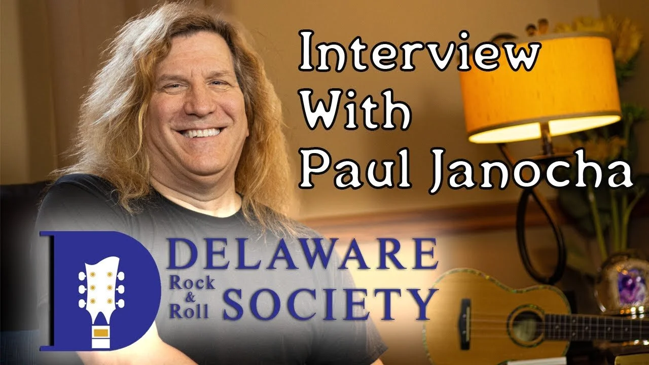 A smiling man with long, wavy blonde hair sitting in a room with a lamp and guitar in the background, with text overlay reading 'Interview with Paul Janocha' and the Delaware Rock & Roll Society logo.