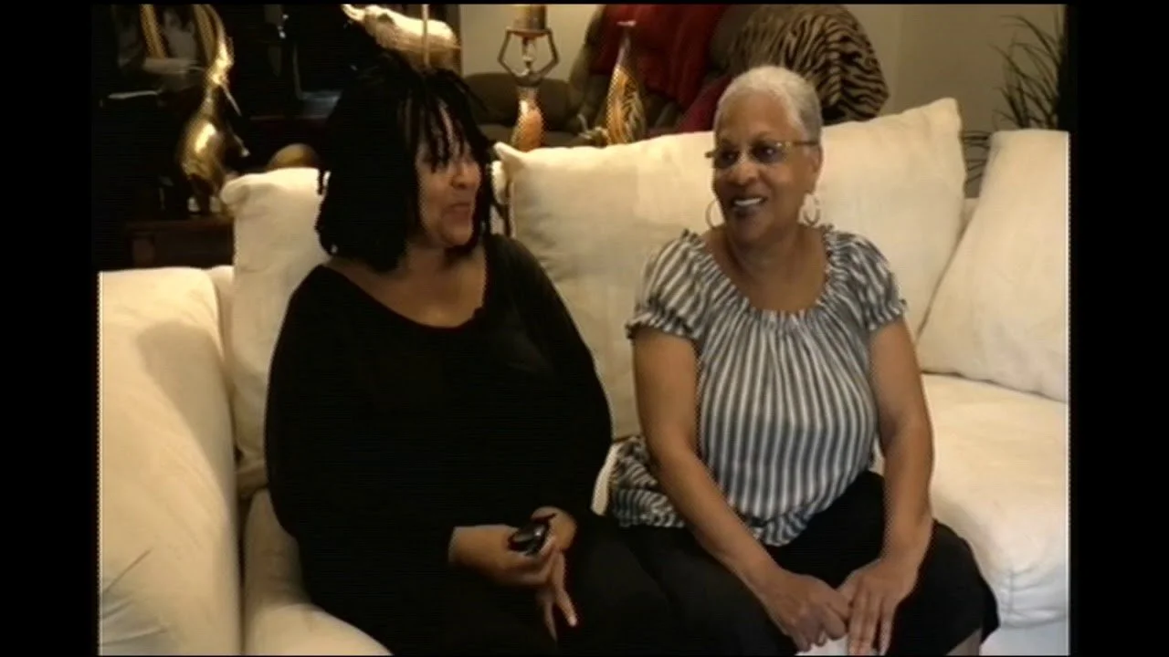 Two women sitting on a cream-colored sofa, engaged in conversation, in a living room with decorative objects and plants in the background.