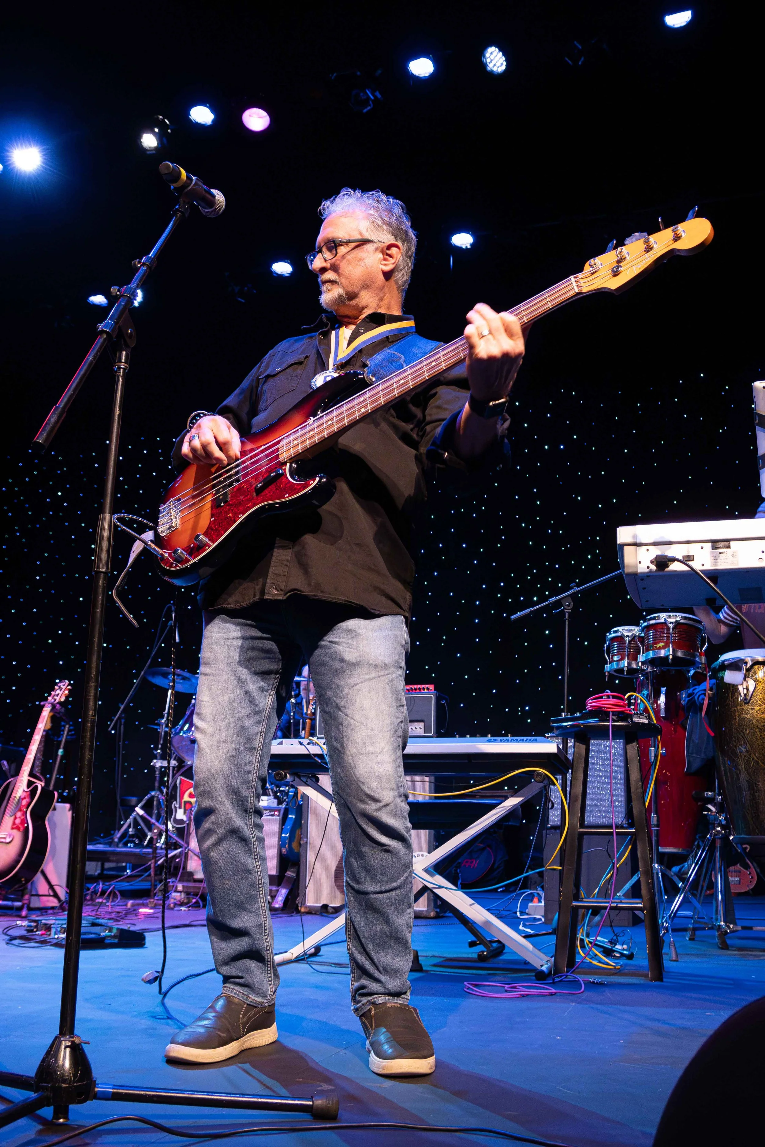A man playing a bass guitar on stage during a concert, with musical instruments and equipment around him and colorful stage lights overhead.