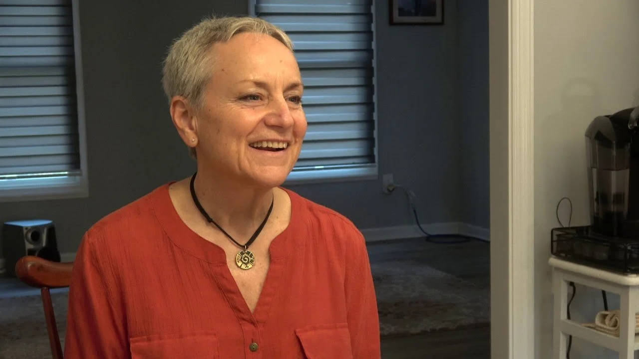 A woman with short gray hair smiling, wearing an orange blouse and a black necklace with a pendant, sitting in a room with windows and blinds.