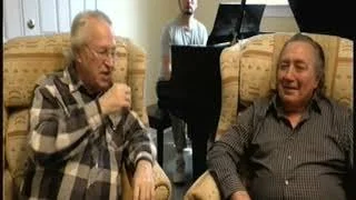 Two older men sitting on a beige sofa, one singing into a microphone and the other smiling. A person playing piano is visible in the background.