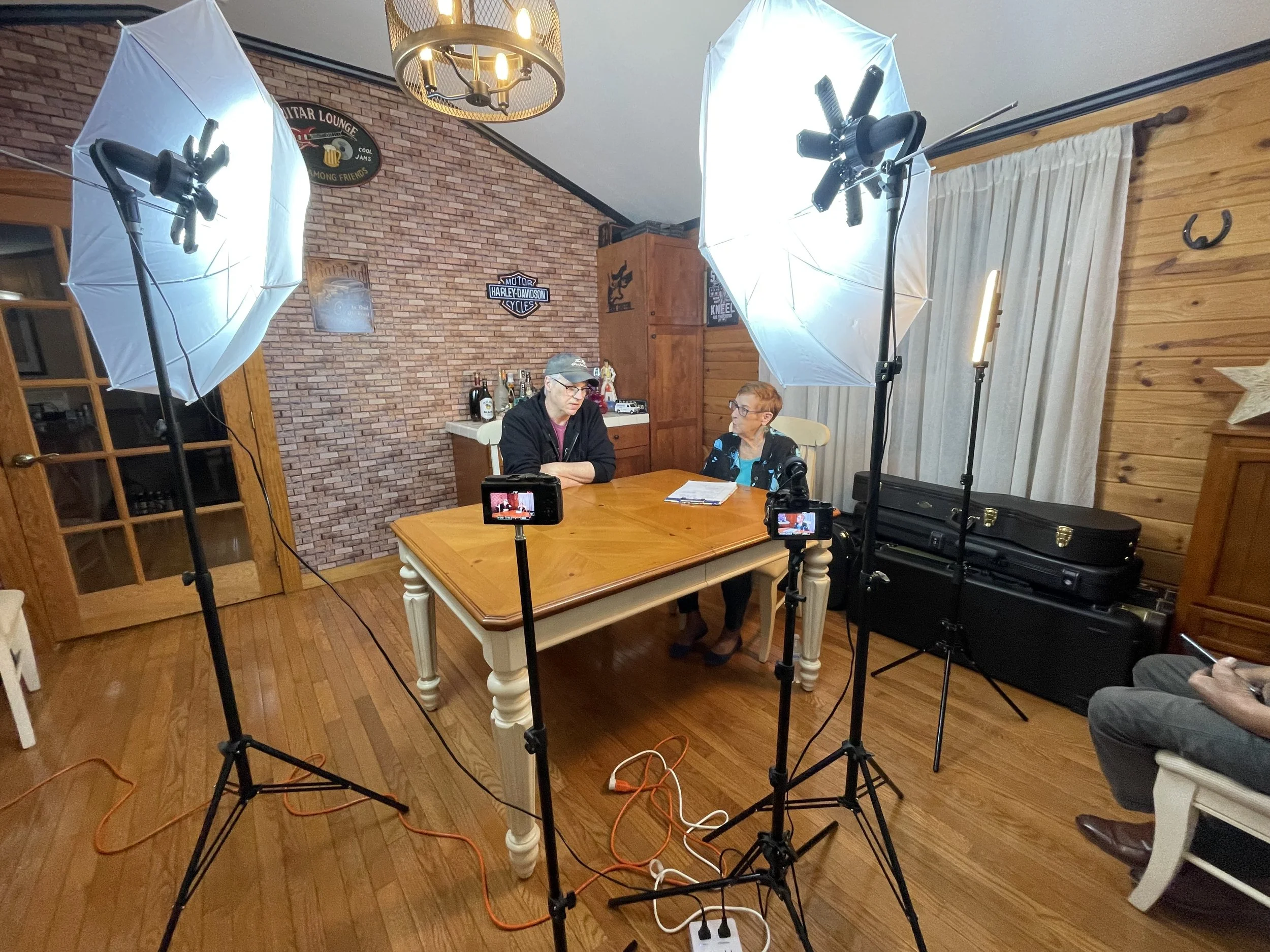Two women seated at a wooden table in a room designed as a home studio, surrounded by professional lighting and camera equipment, with a brick wall and wooden paneling in the background.