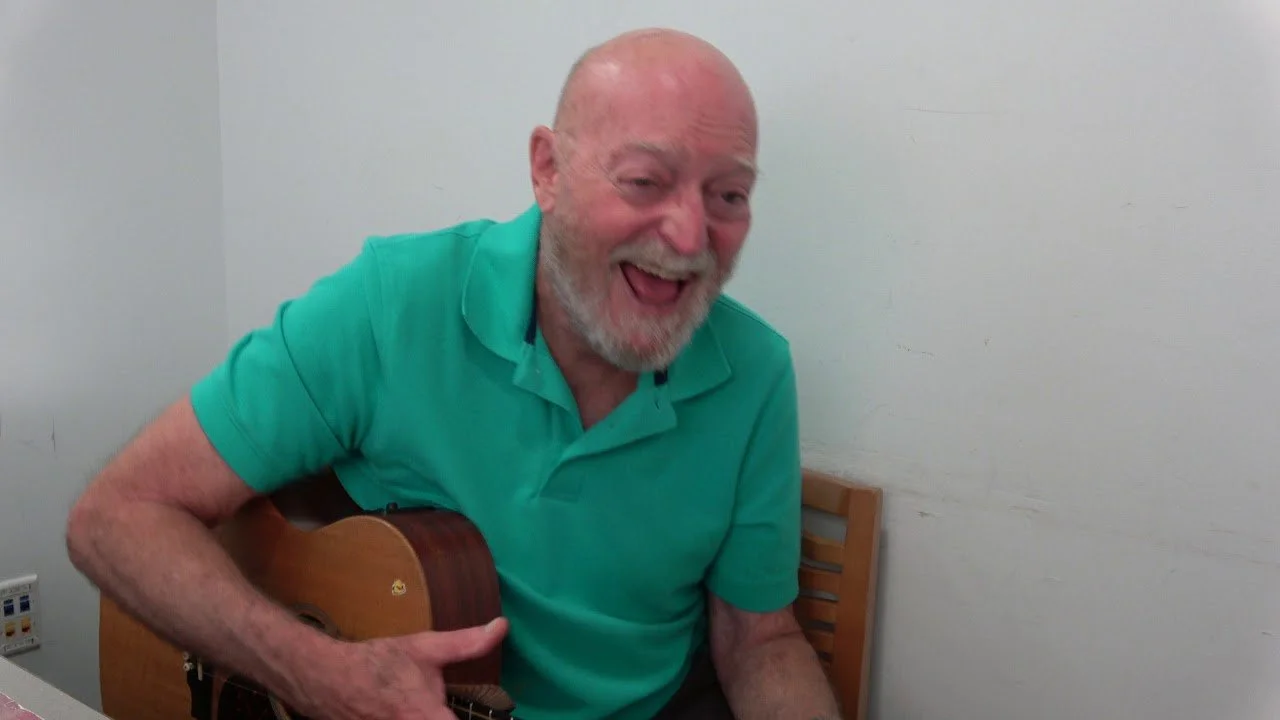 An elderly man with a bald head, gray beard, and mustache, wearing a turquoise polo shirt, sitting at a table, playing an acoustic guitar, and smiling happily.