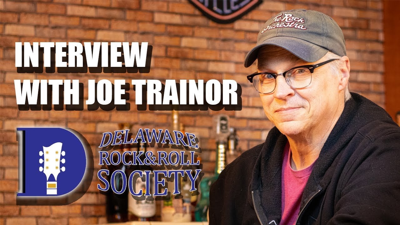 A man wearing glasses, a gray cap with the logo of a motorcycle club, and a black hoodie, sitting in front of a brick wall. The image features text that reads "Interview with Joe Trainor" and an emblem of the Delaware Rock & Roll Society.