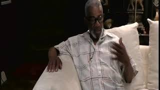 Older man sitting on a bed in a bedroom, wearing glasses and a light-colored, checkered shirt, with his hand on his chest.