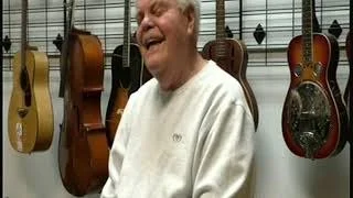 An older man smiling and laughing in a music store, surrounded by hanging guitars.