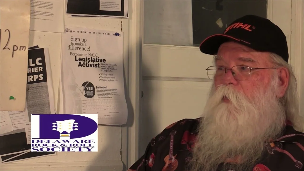 An elderly man with a long white beard, glasses, wearing a black cap and a shirt with guitar graphics, sitting in front of a notice board with flyers and papers, including one titled 'Sign up to make a difference! Become an NALC Legislative Activist.' The Delaware Rock & Roll Society logo is visible in the bottom left corner.