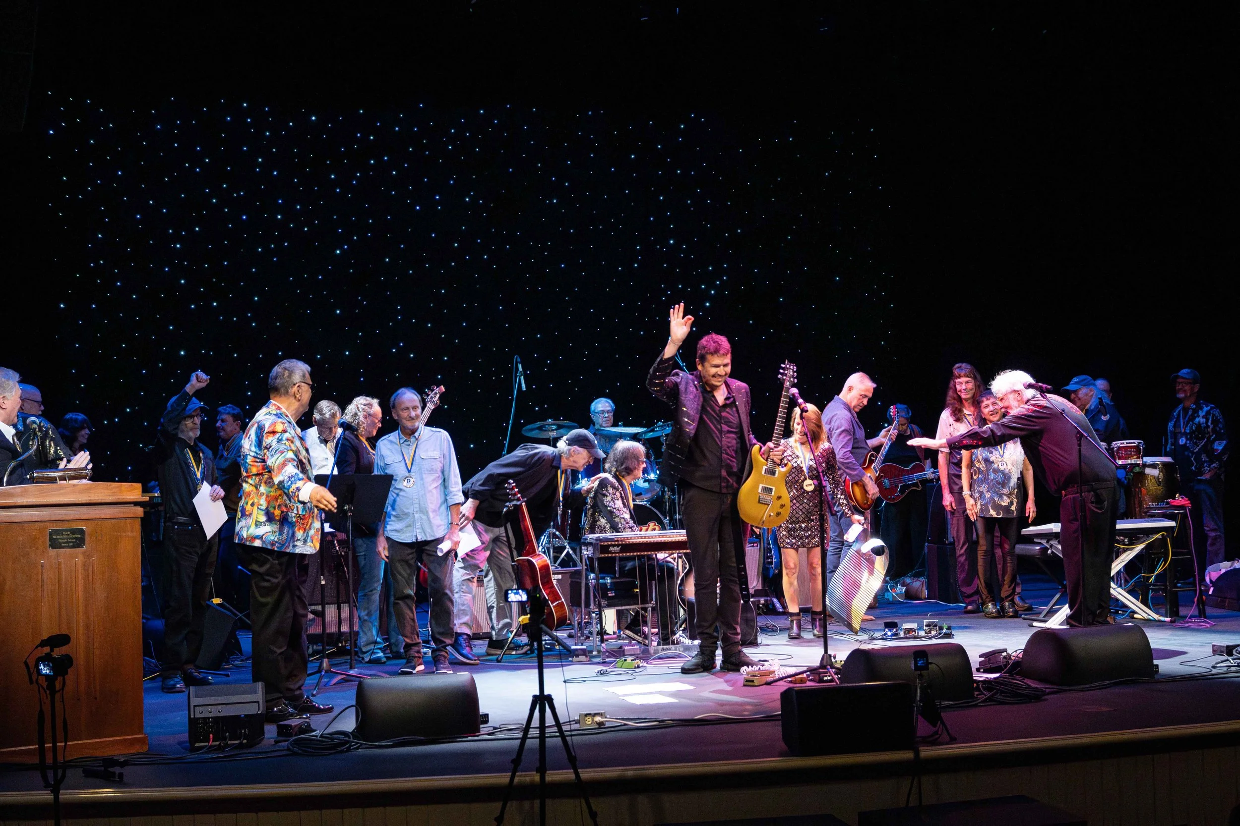 Group of musicians and performers on stage, some holding instruments, with a dark backdrop featuring small, bright lights, during a live music event.