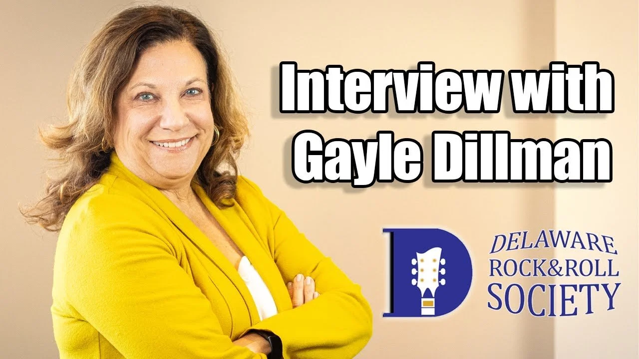 A woman with wavy brown hair wearing a yellow blazer smiling with arms crossed, standing next to a logo of the Delaware Rock & Roll Society with a guitar illustration.