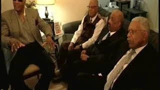 Four men are seated in a room having a discussion. One man is standing and speaking, while the other three listen. The setting appears to be informal or professional, with a couch and lamp in the background.