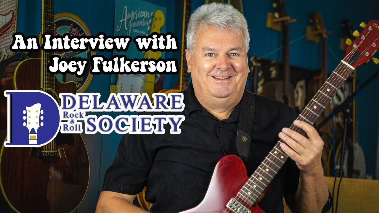 Man with gray hair and a black shirt playing a red electric guitar, smiling, with musical instruments in the background, including guitars and a blue sign that reads Mardi Gras.