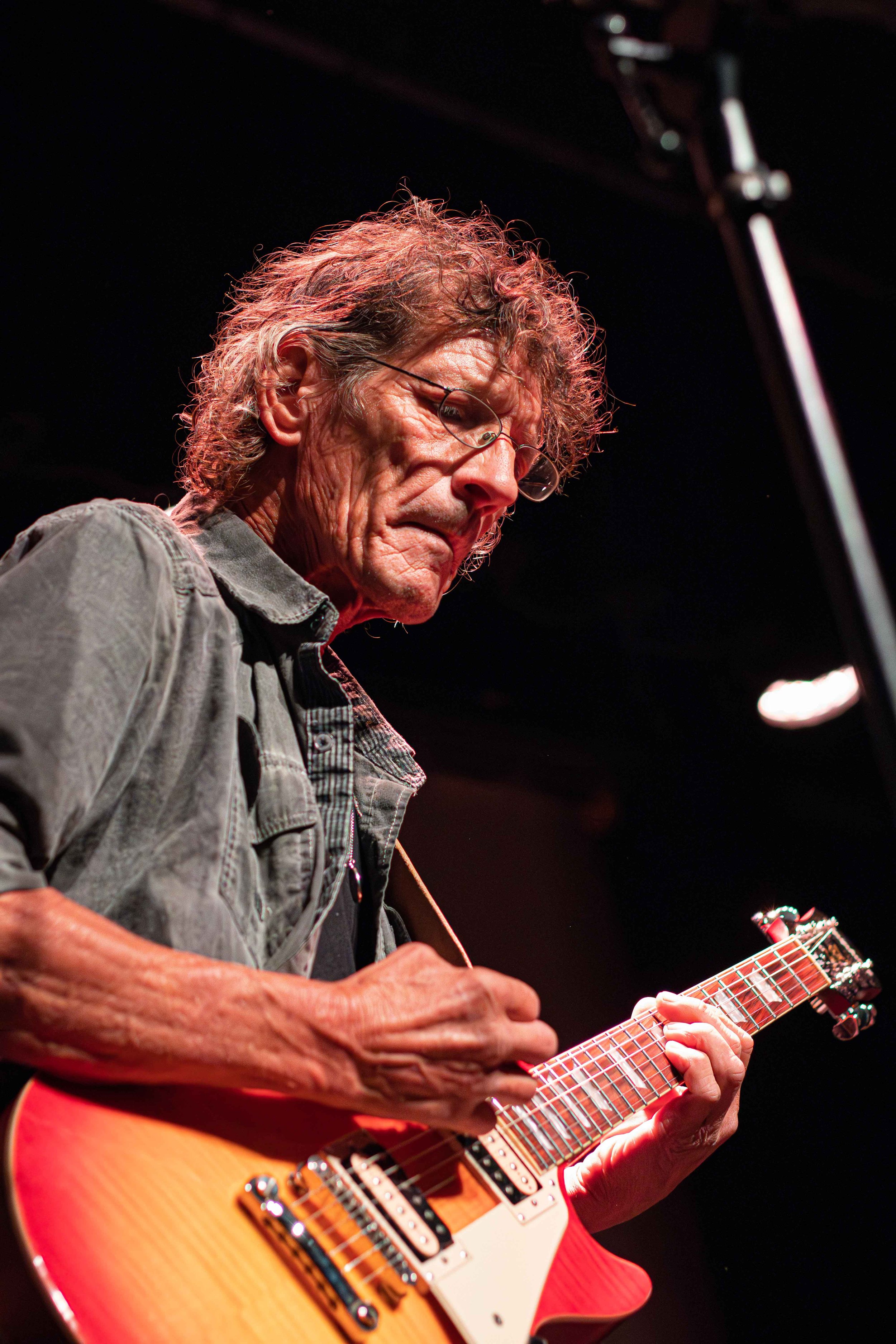 An elderly man with curly gray hair and glasses playing an electric guitar on stage.