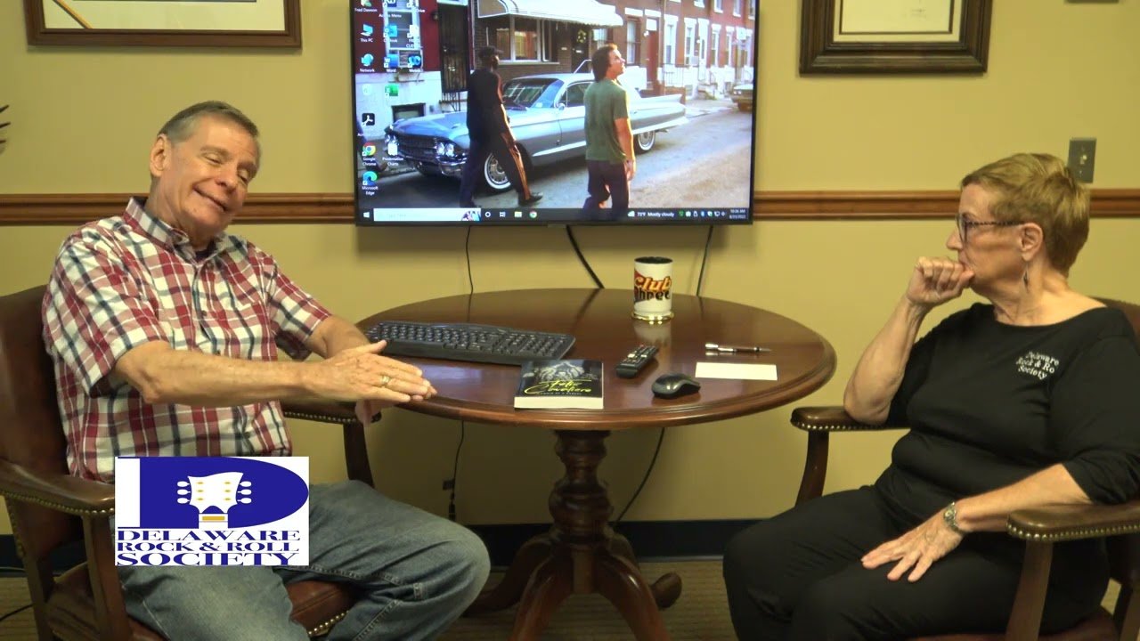 A man and a woman sit at a round wooden table in a room, engaged in conversation. The man is wearing a checkered shirt and smiling, while the woman is wearing glasses and a dark shirt, appearing thoughtful. On the table are a computer keyboard, a remote, a notebook, a pen, a cup with a logo, and a TV remote. Behind them, a large TV screen displays a scene with two people and a classic car. There is a logo in the bottom left corner that reads 'Delaware Rock & Roll Society.'