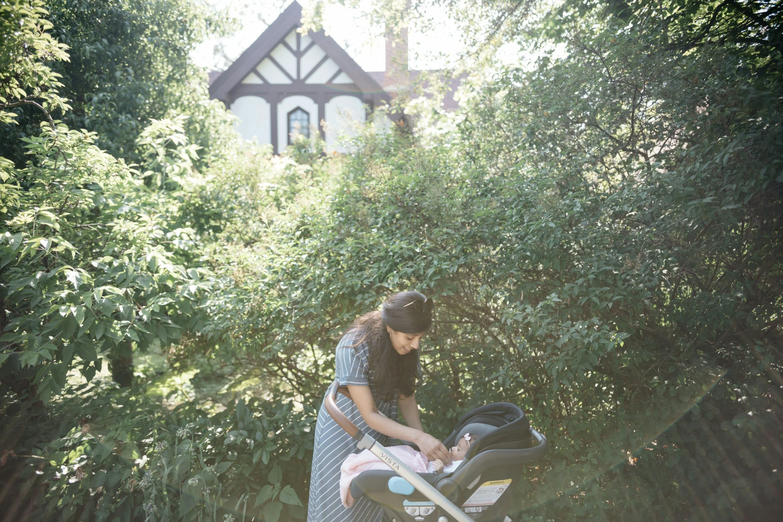 A woman with long dark hair and a striped dress is smiling and leaning over a baby in a car seat stroller outdoors surrounded by green trees and bushes.