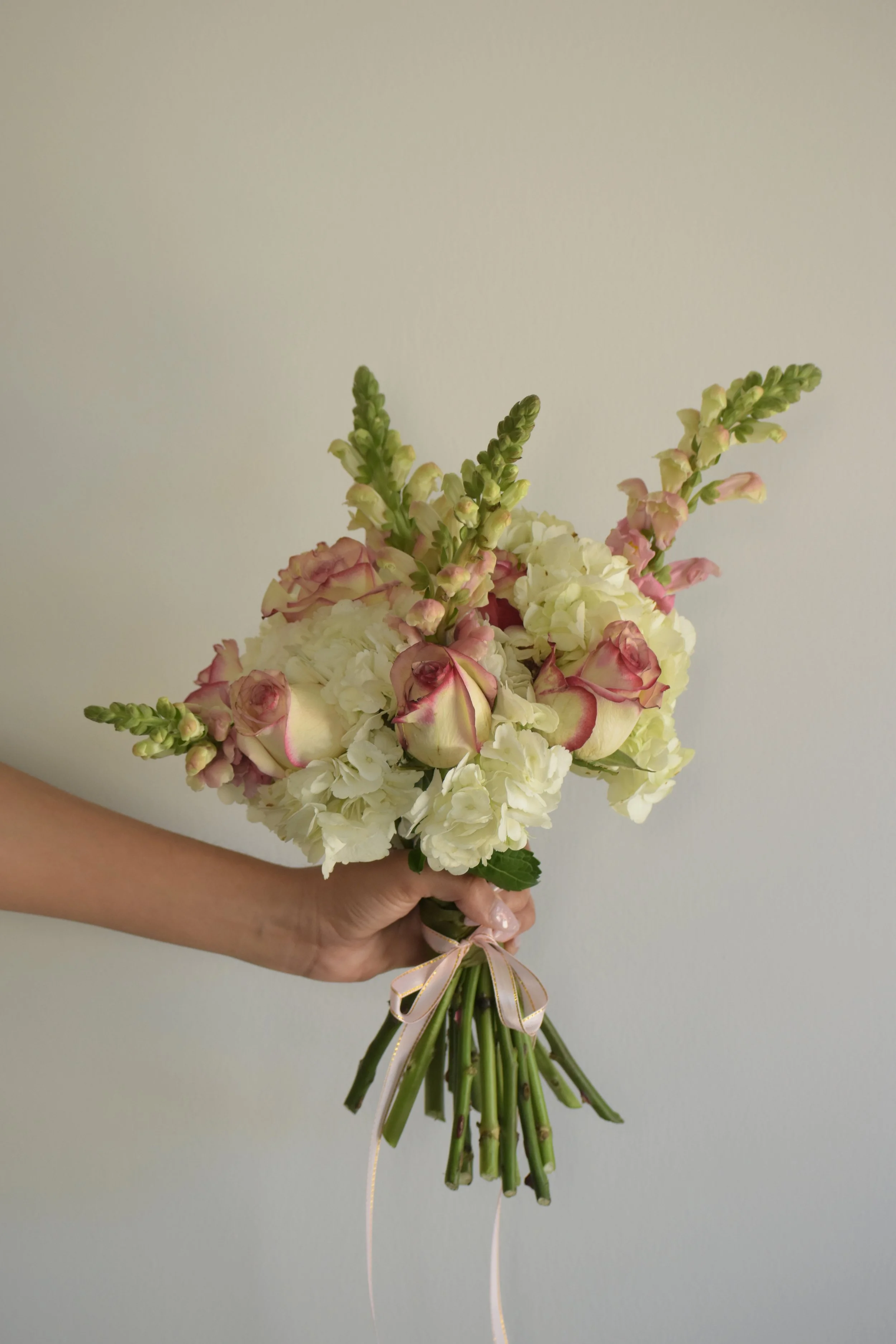 A hand holding a bouquet of white and pink flowers, tied with a pink ribbon, against a plain white background.