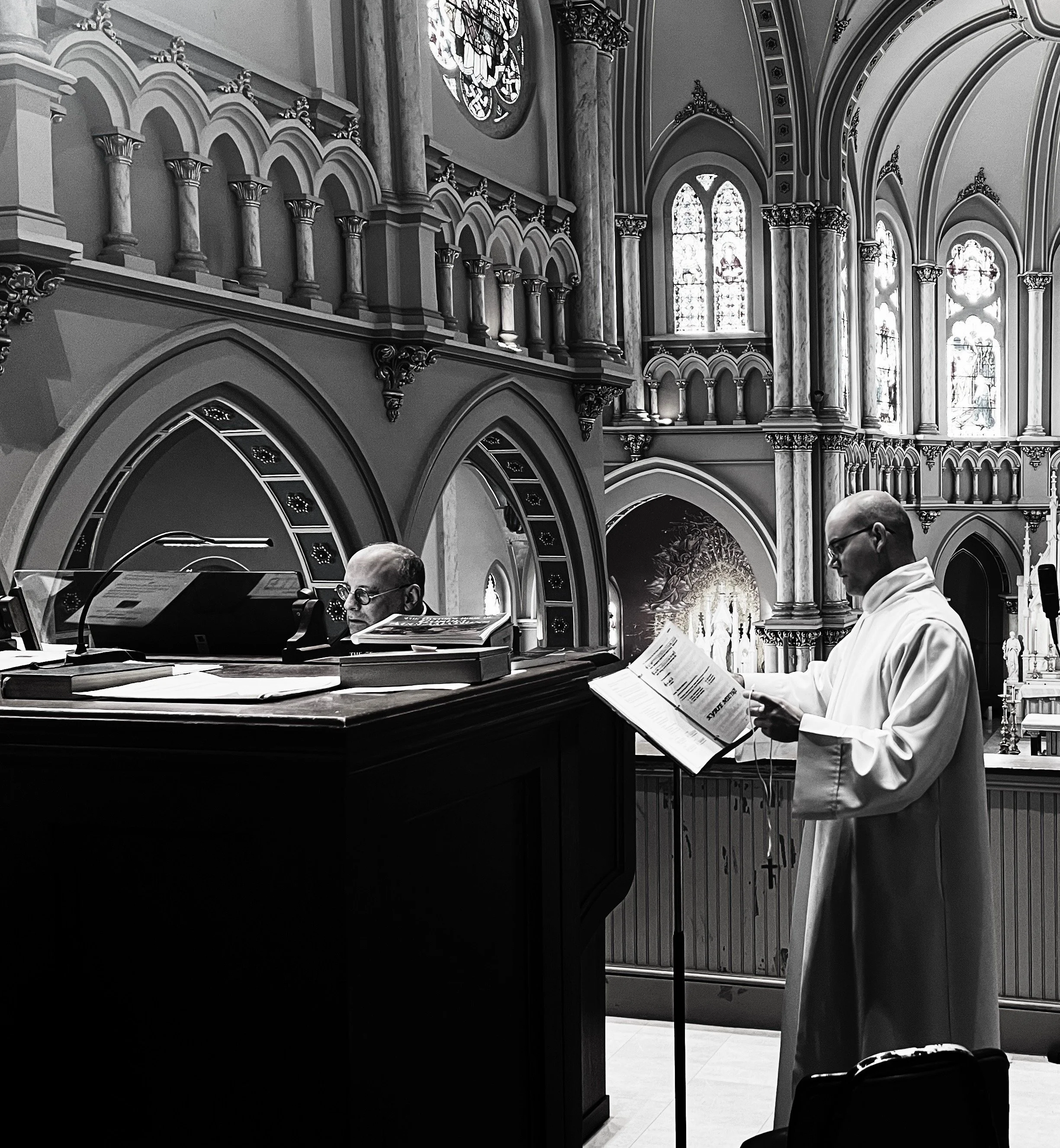 A black-and-white photo inside a church showing a priest reading from a book and another man playing a keyboard, with ornate architecture and stained glass windows in the background.