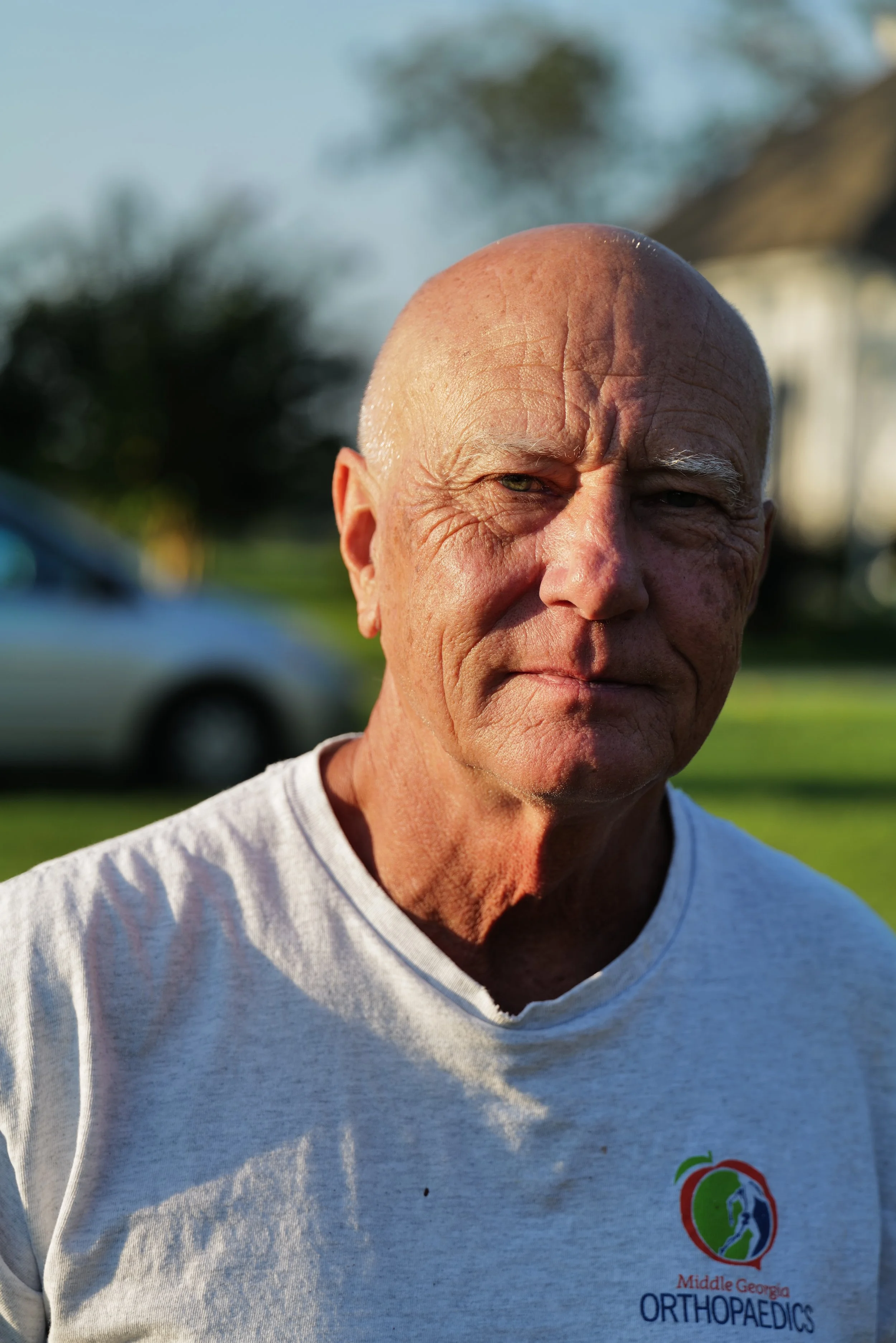 A close-up of an elderly man with a bald head outdoors, wearing a gray t-shirt with a 'Middle Georgia Orthopaedics' logo, with a car and house in the background during daytime.