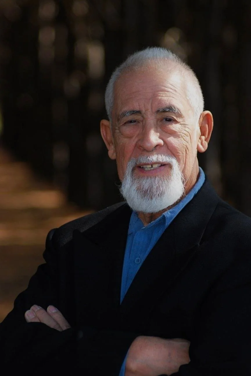 An elderly man with a white beard and mustache, smiling, wearing a black jacket and a blue shirt, standing outdoors with a blurred background.