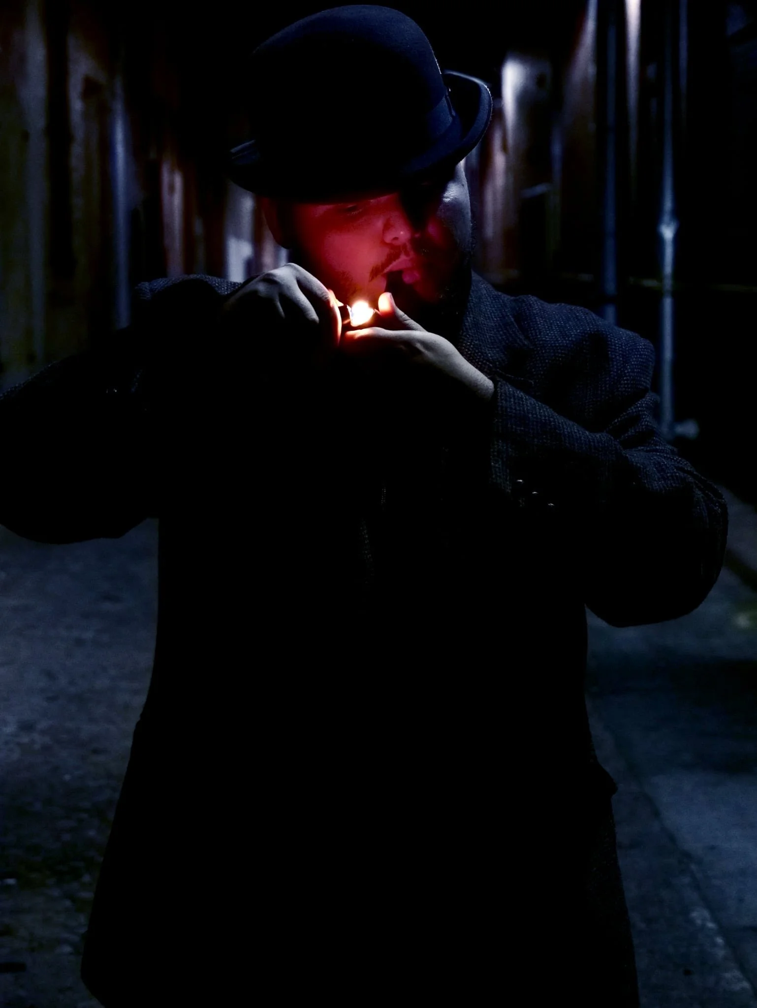 Man wearing a black hat and coat lighting a cigarette in a dimly lit alley.