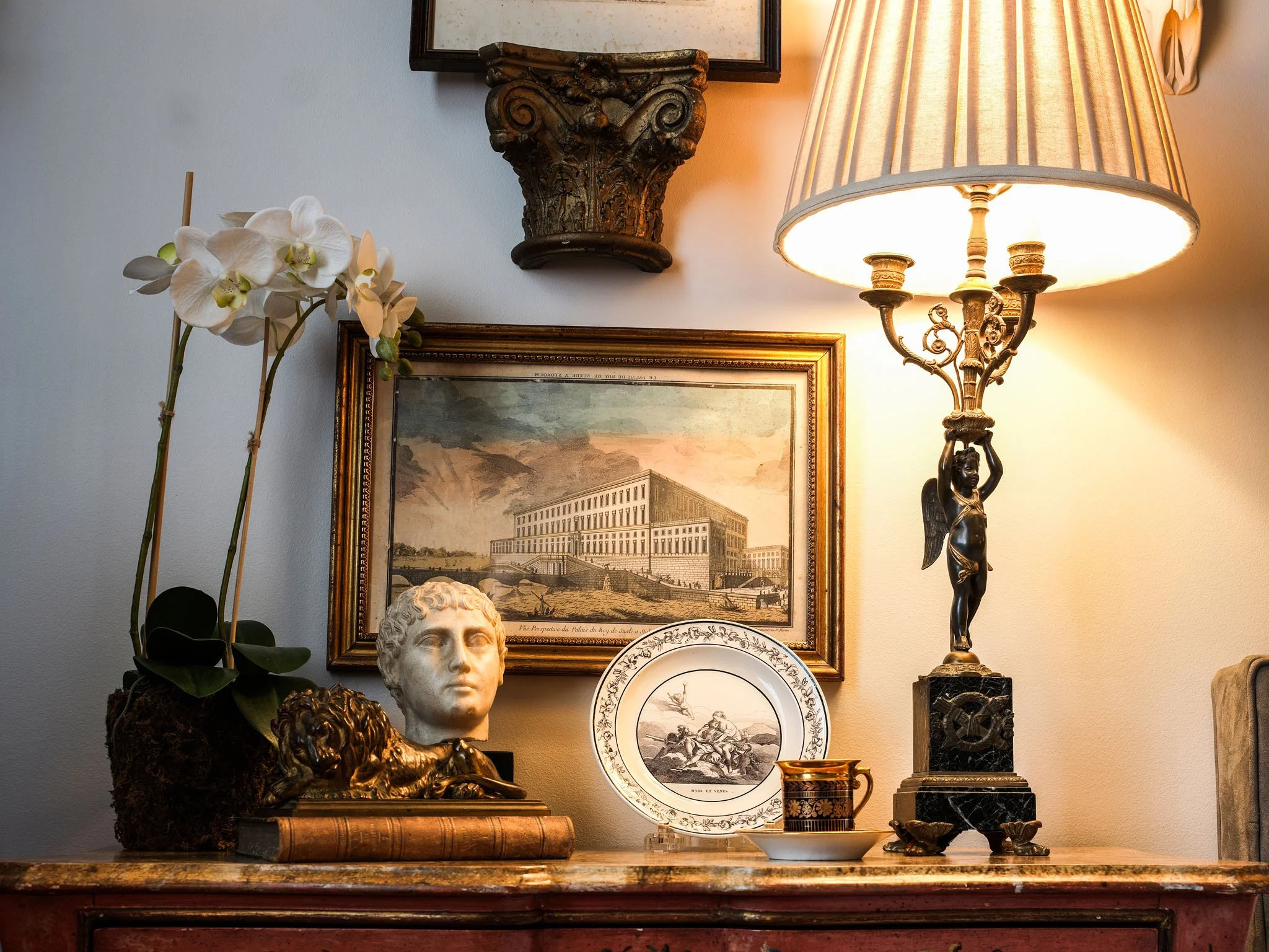 A vintage interior decor display on a wooden surface, featuring orchids, a carved bust, a lion sculpture, framed artwork, a decorative plate, a cup and saucer, and a decorative table lamp with an angel figurine base, set against a wall with additiona