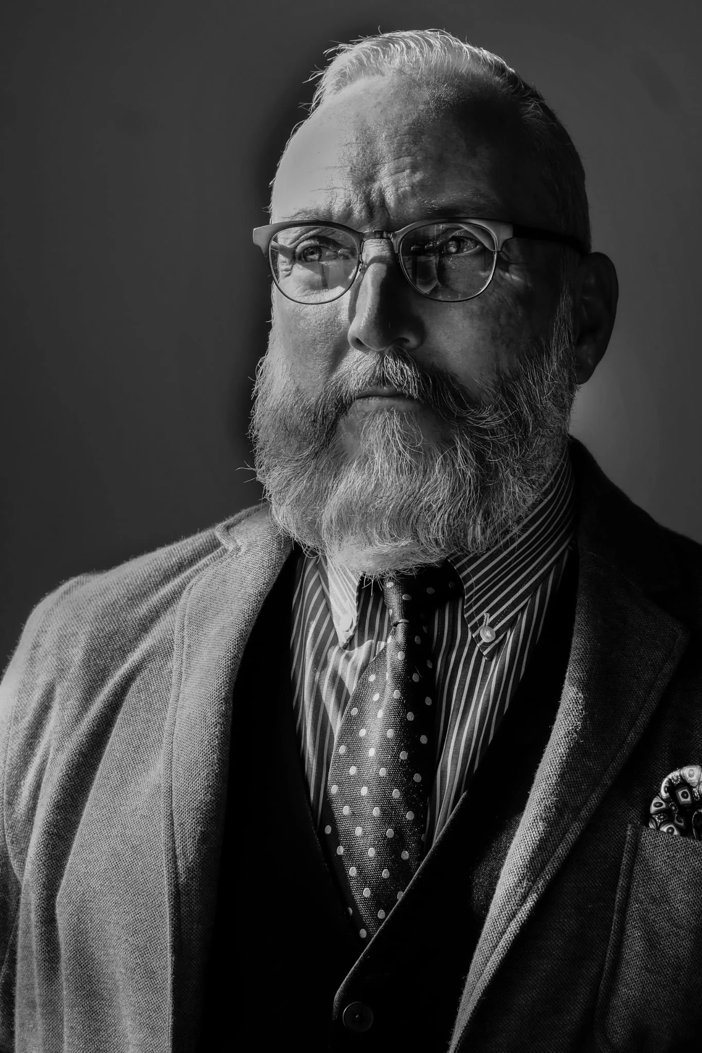 Black and white portrait of a man with glasses, a beard, and mustache, wearing a suit, striped shirt, tie, and pocket square, looking thoughtfully to the side.