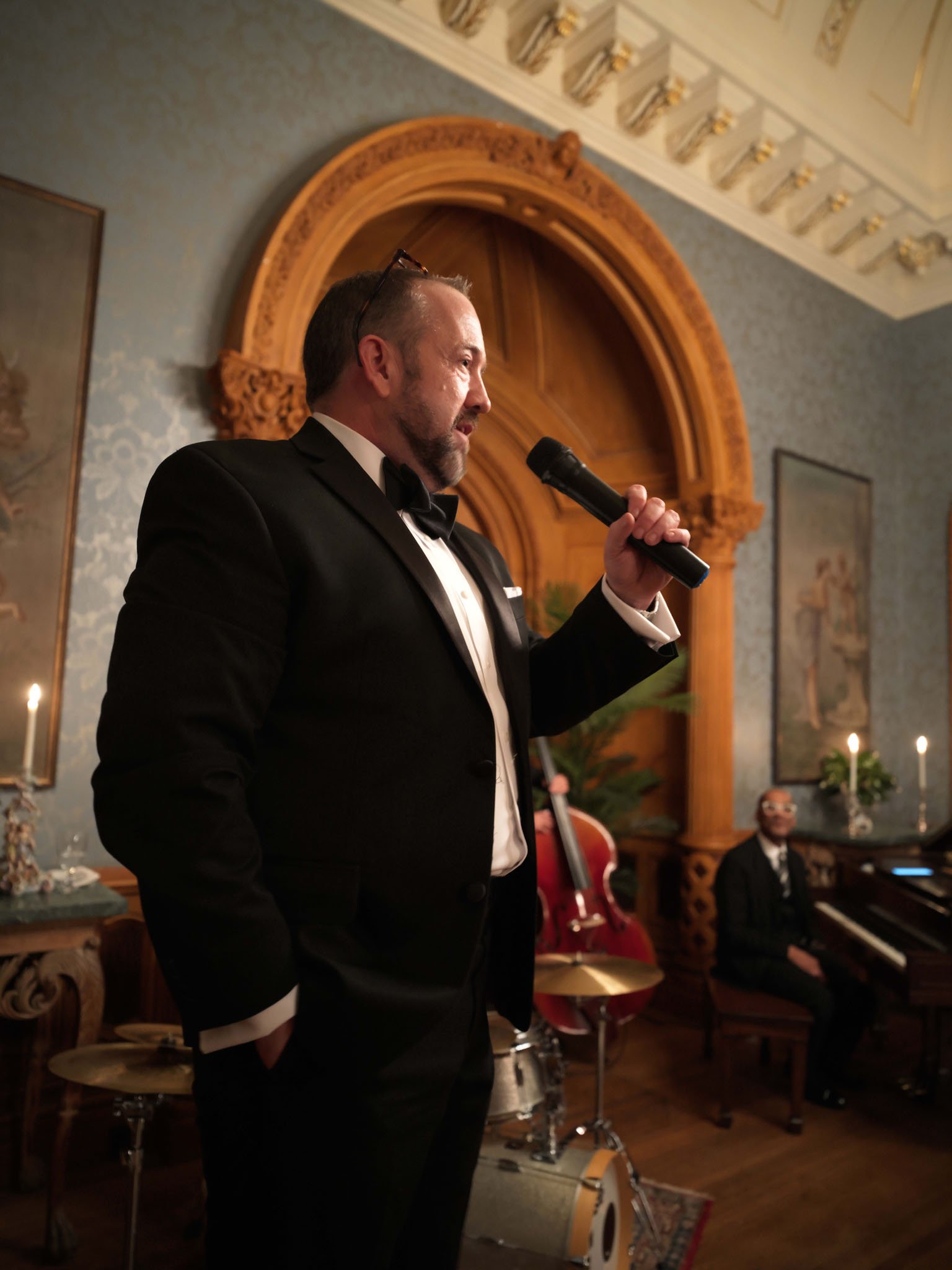 A man in a black tuxedo holding a microphone, singing or speaking at an indoor event. A musician with sunglasses is sitting at a piano in the background.
