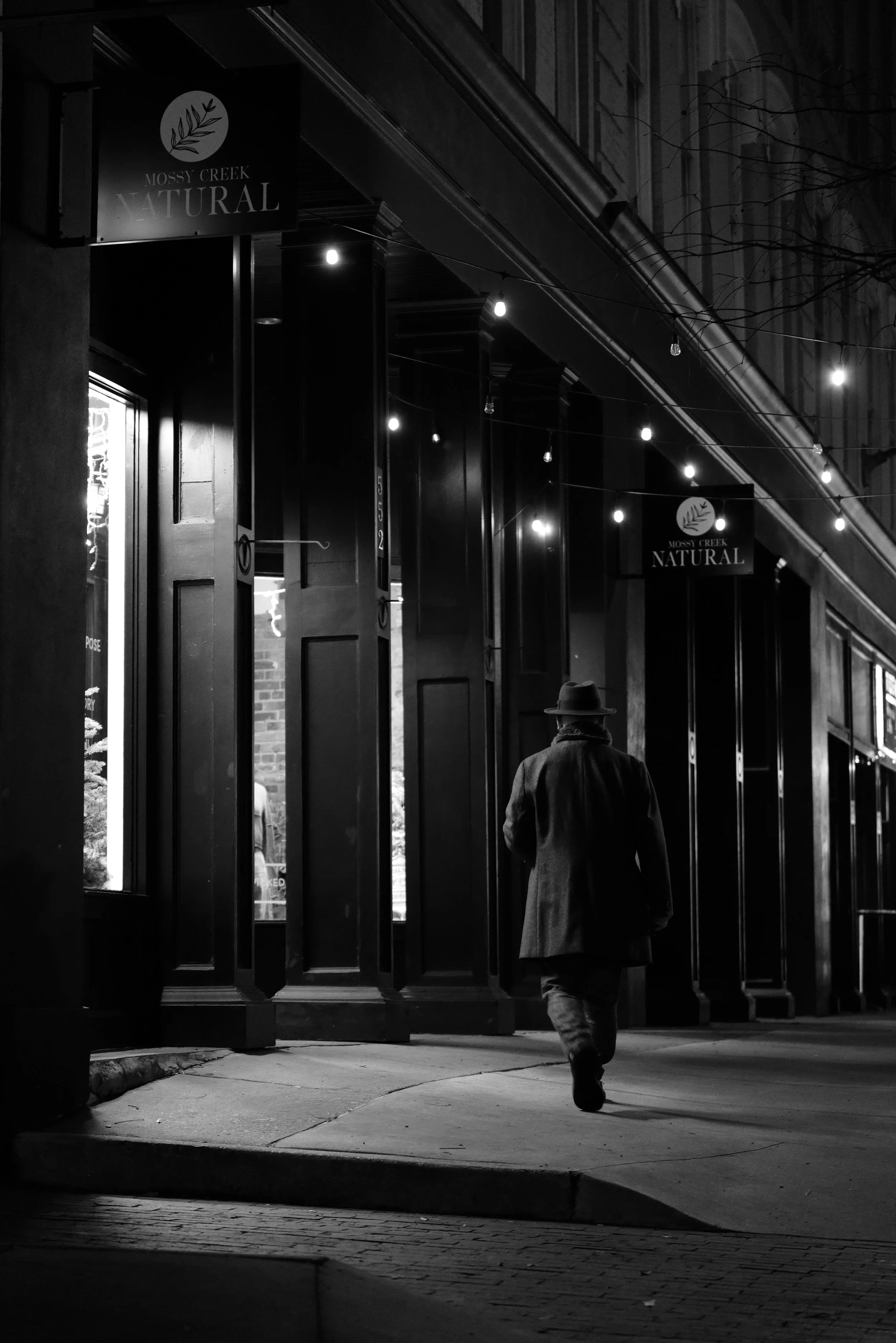 A person walking on the sidewalk at night near storefronts with illuminated signs, black and white photo.