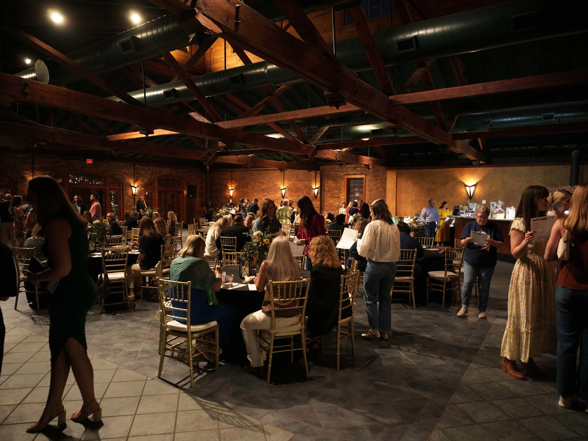 People gathering at a social event in a spacious indoor venue with exposed brick walls, wooden beams, and warm lighting. Guests are seated at round tables and standing, engaging in conversations, with a bar or service area in the background.