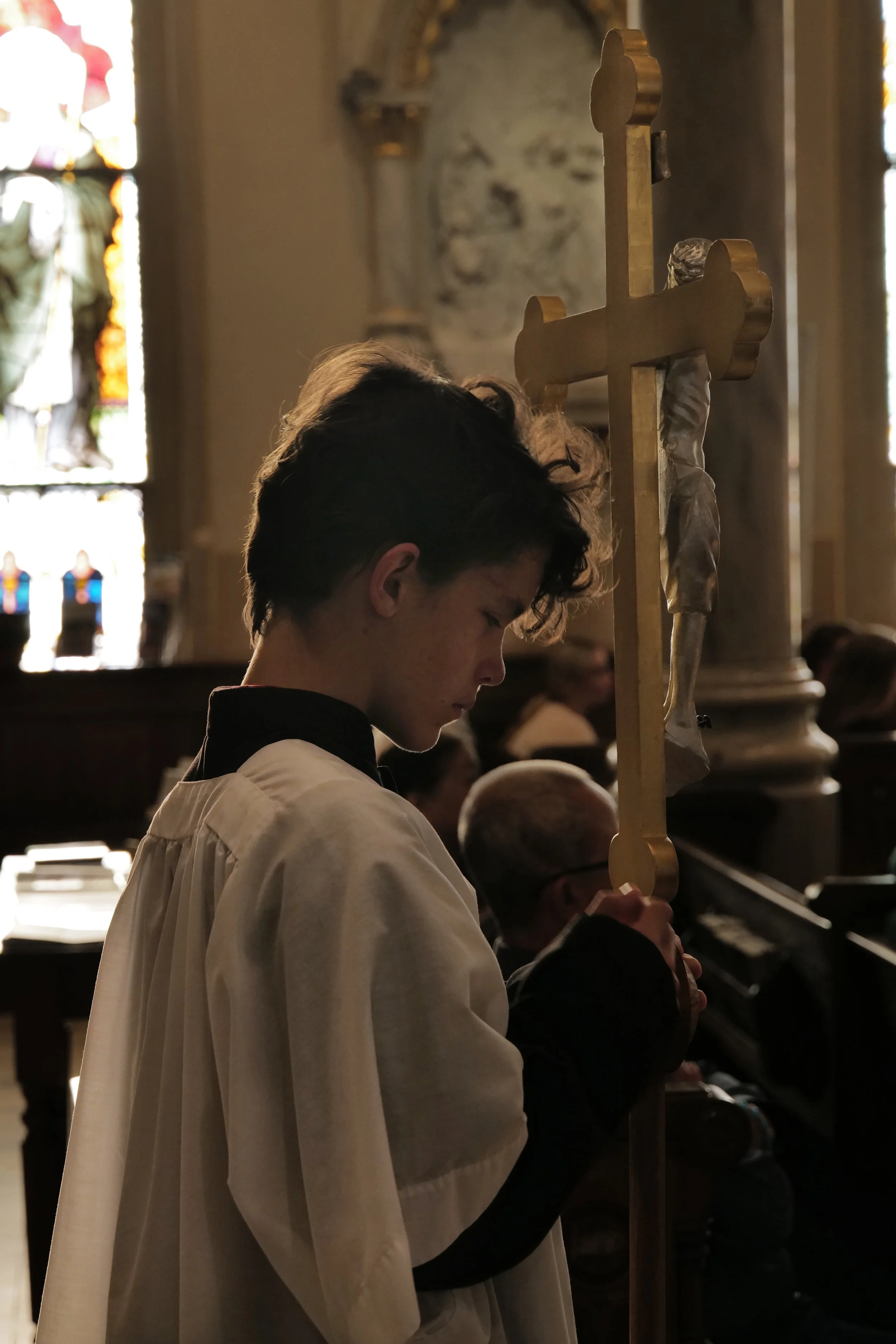 A young person in church robes holding a processional cross, standing solemnly during a religious service inside a church with stained glass windows.