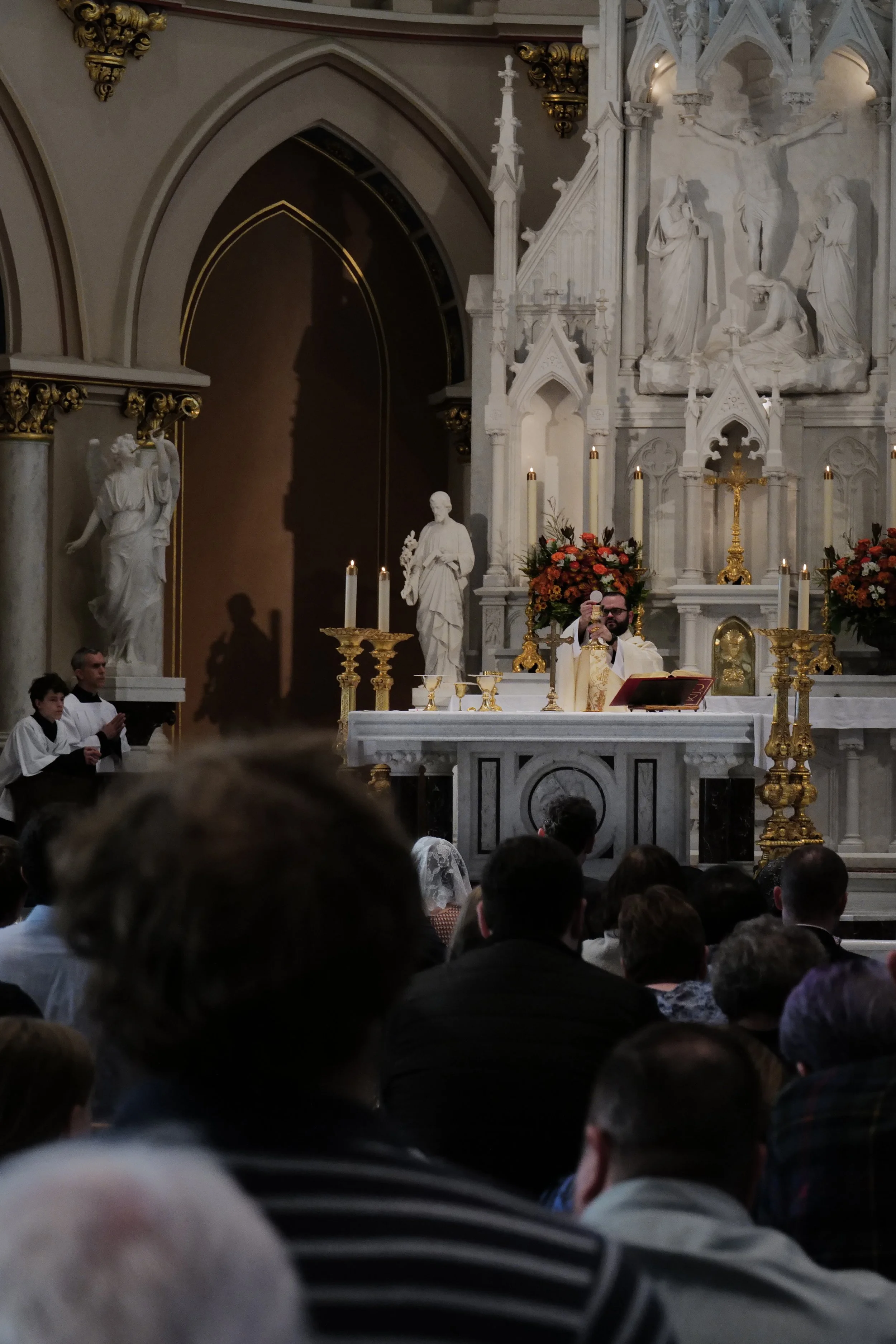 A Catholic priest at the altar celebrating mass in a church with ornate statues and religious iconography. Congregation seated in the pews.