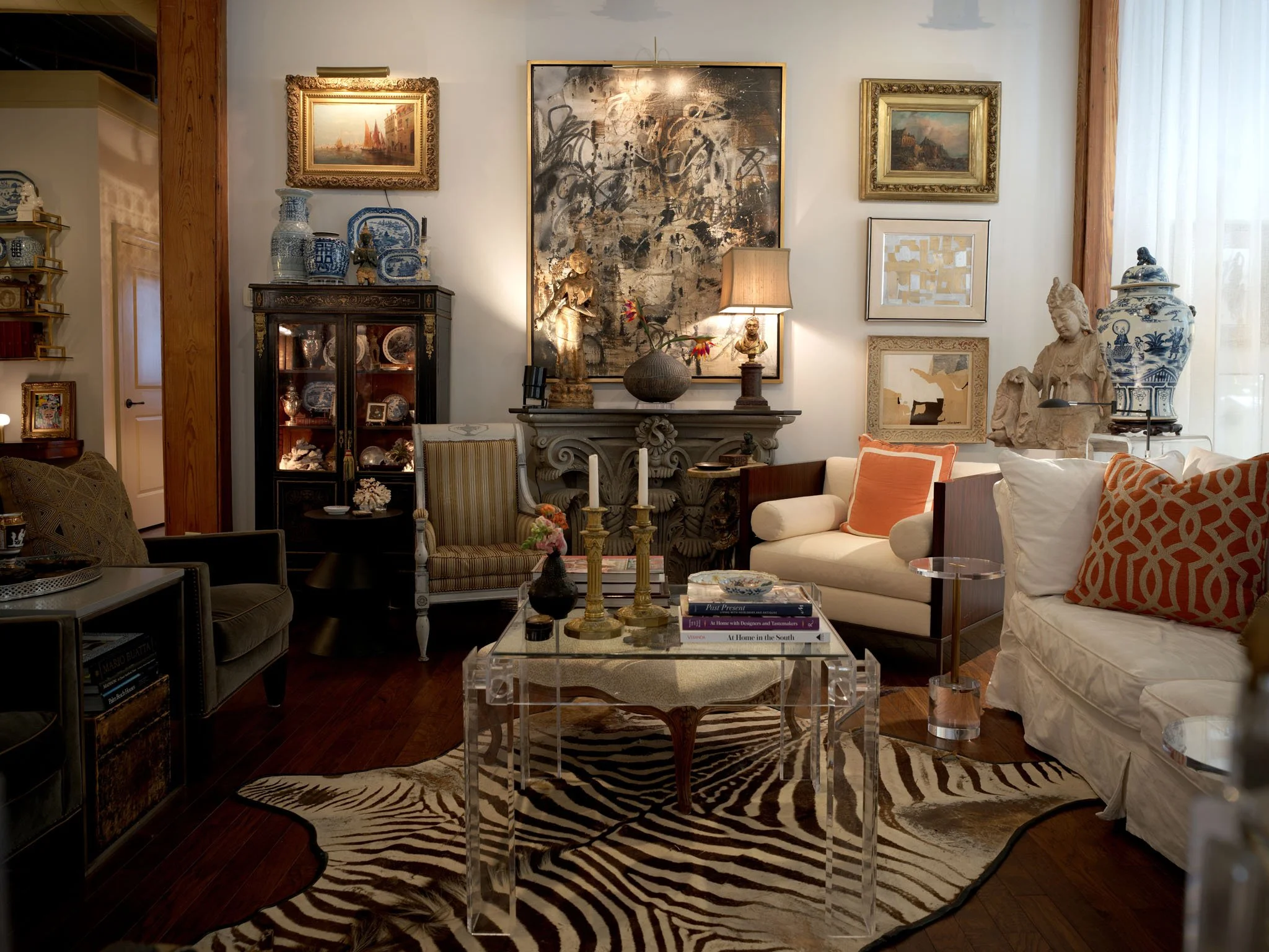 Living room with antique and modern decor, featuring a zebra print rug, glass coffee table with candles, sofa with orange patterned pillow, artwork on the walls, and decorative vases and sculptures.