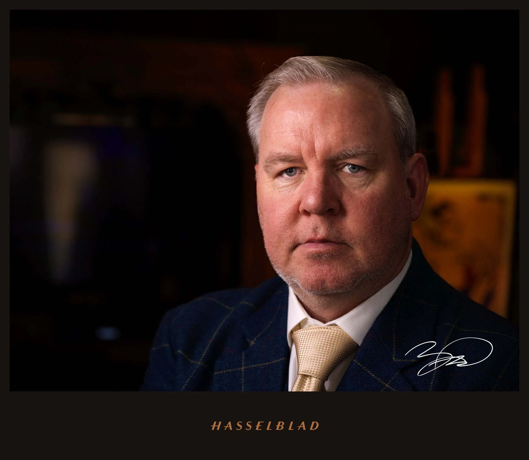 Close-up of a middle-aged man with gray hair wearing a dark checkered suit, white shirt, and beige tie, looking serious, signature in the lower right corner, background with dark wooden elements, and the word 'HASSELBLAD' at the bottom.