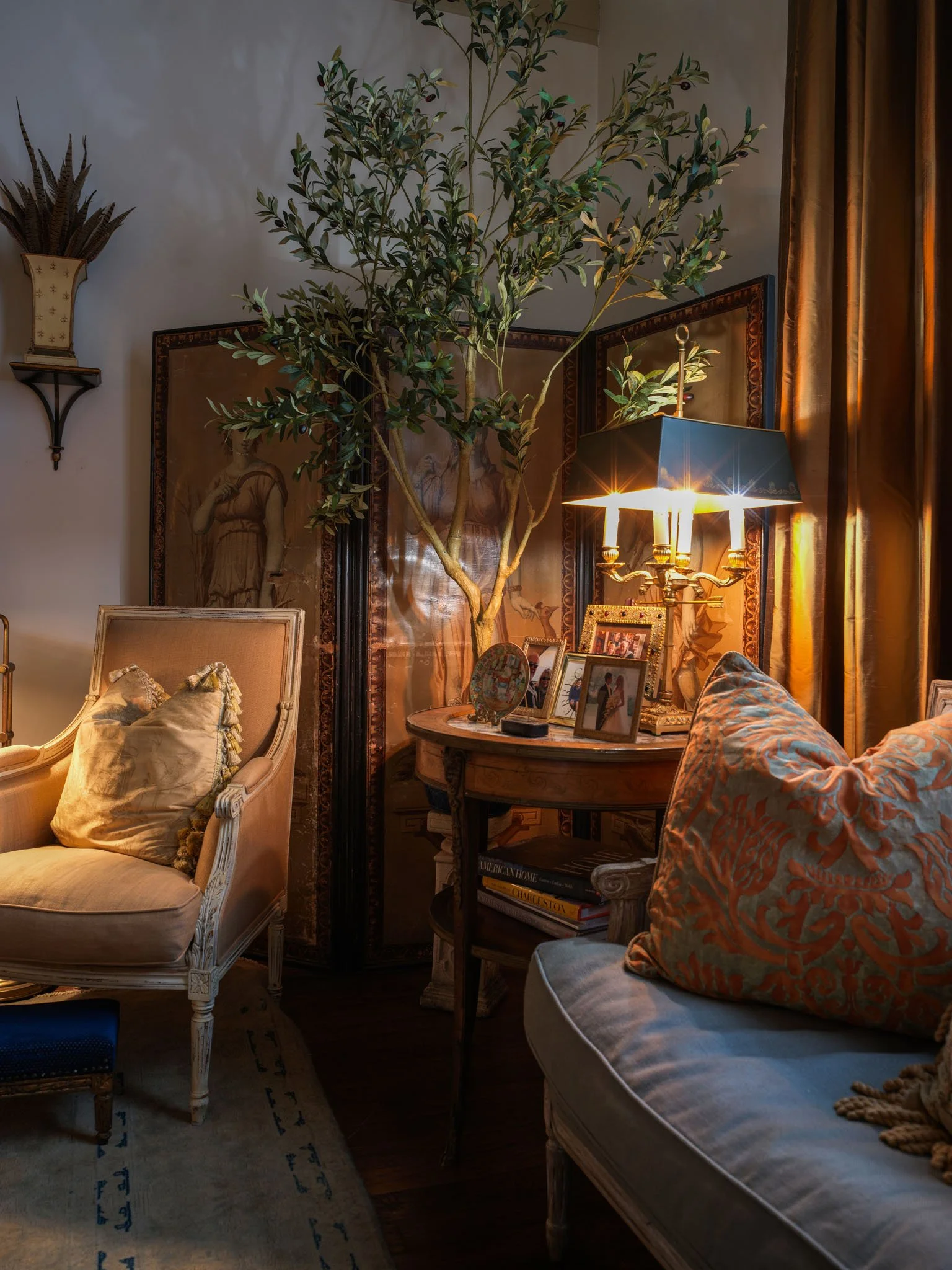 A cozy living room corner with antique furniture, a large potted plant, and framed photos on a small round table illuminated by a chandelier lamp. The room features warm lighting, draped gold curtains, and decorative wall art.