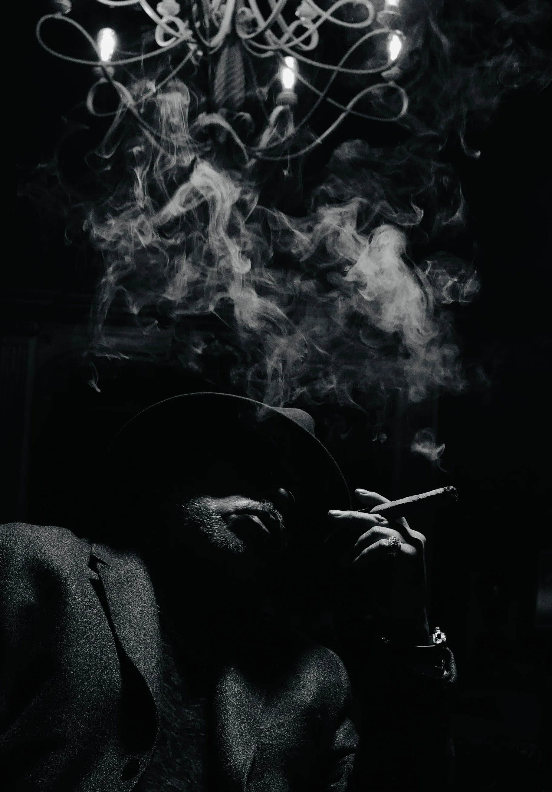 A person wearing a hat and suit, sitting in a dark room, smoking a cigar with smoke rising around them, illuminated by a chandelier.