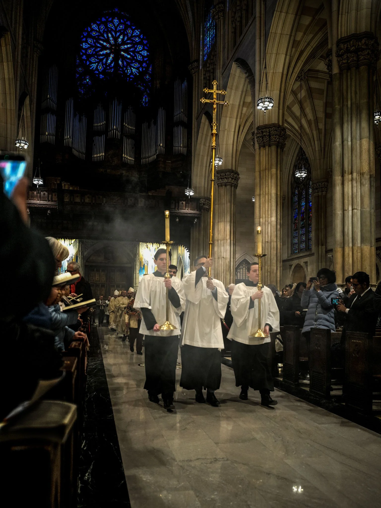 Young men in white robes and black cassocks carrying candles and a processional cross inside a Gothic cathedral during a religious service.