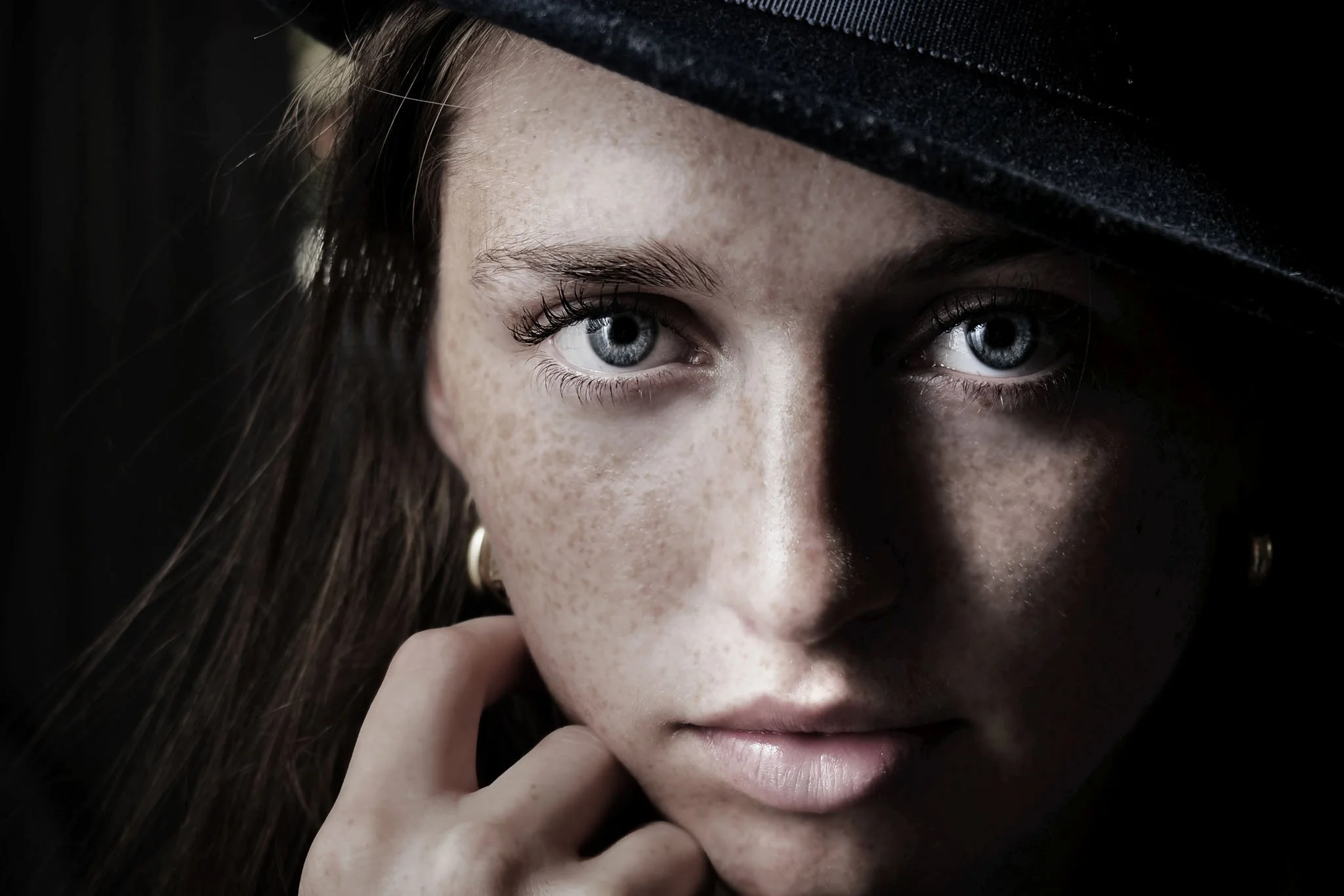 Close-up of a young woman with blue eyes, freckles, and long brown hair, wearing a black hat tilted over her forehead, touching her chin with her hand.