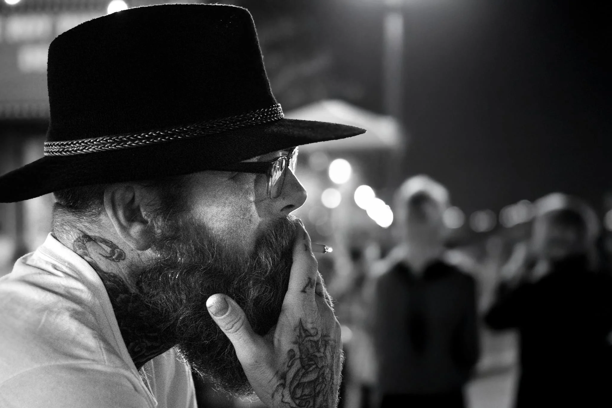 A man with a beard and tattoos, wearing glasses and a wide-brimmed hat with a decorative band, is smoking and resting his head on his hand in a crowded outdoor setting at night.