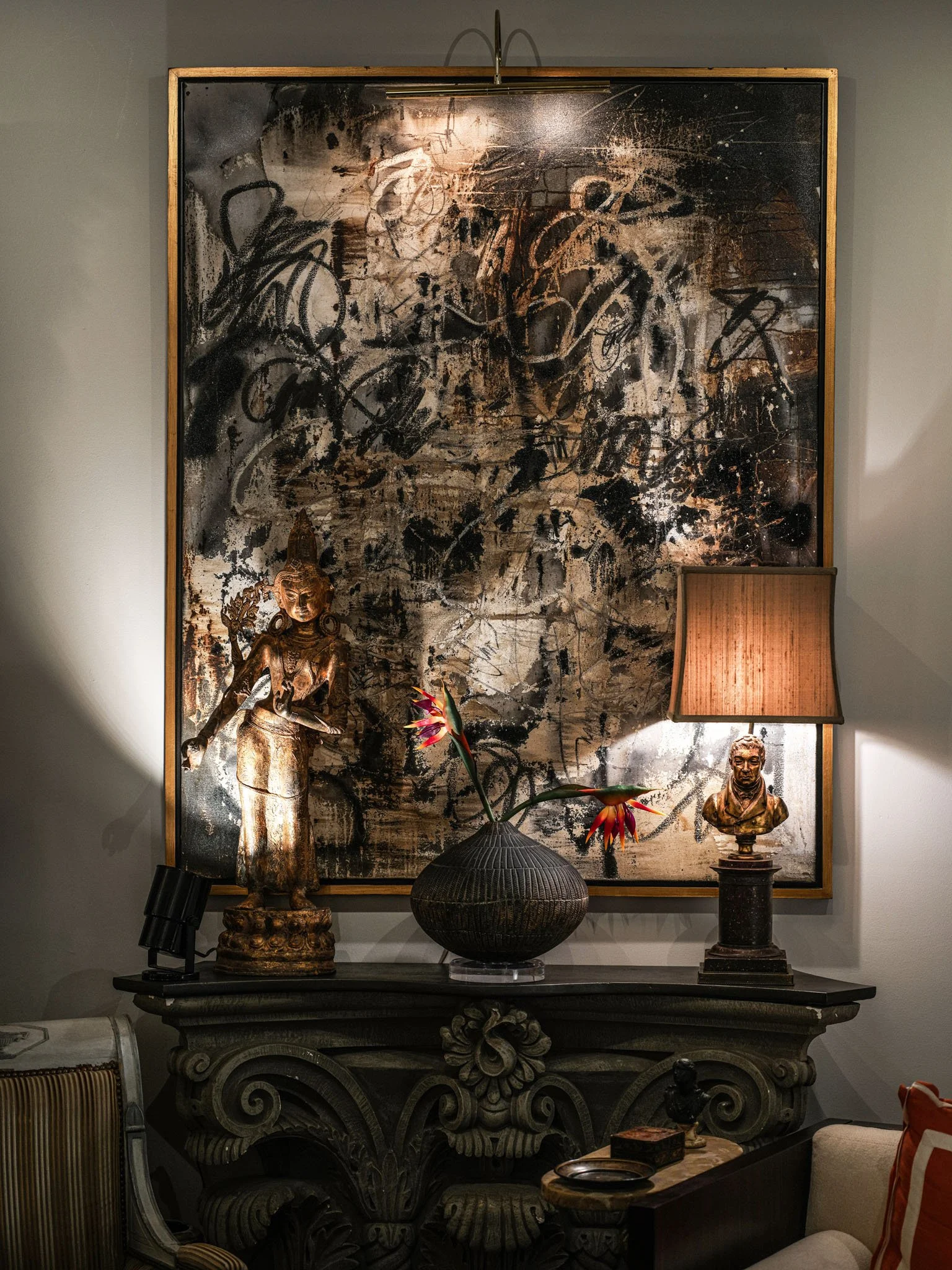 Decorative interior with a large abstract painting, a statue of a woman, a flower vase with pink flowers, a bust sculpture, a lit table lamp, and a carved console table.